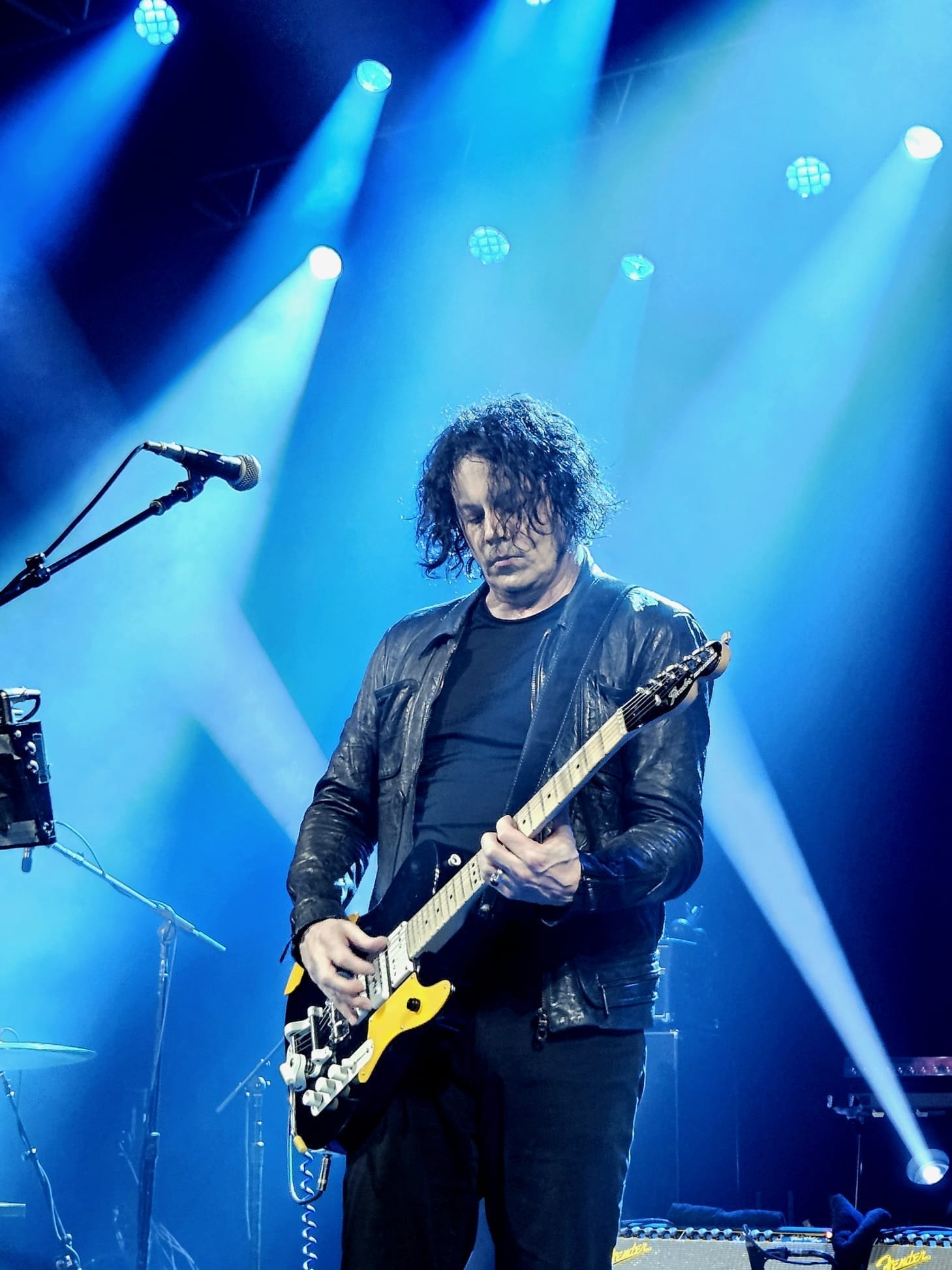 Jack White plays a black and yellow guitar under blue stage lighting, wearing a black leather jacket, with his dark curly hair falling over his face.