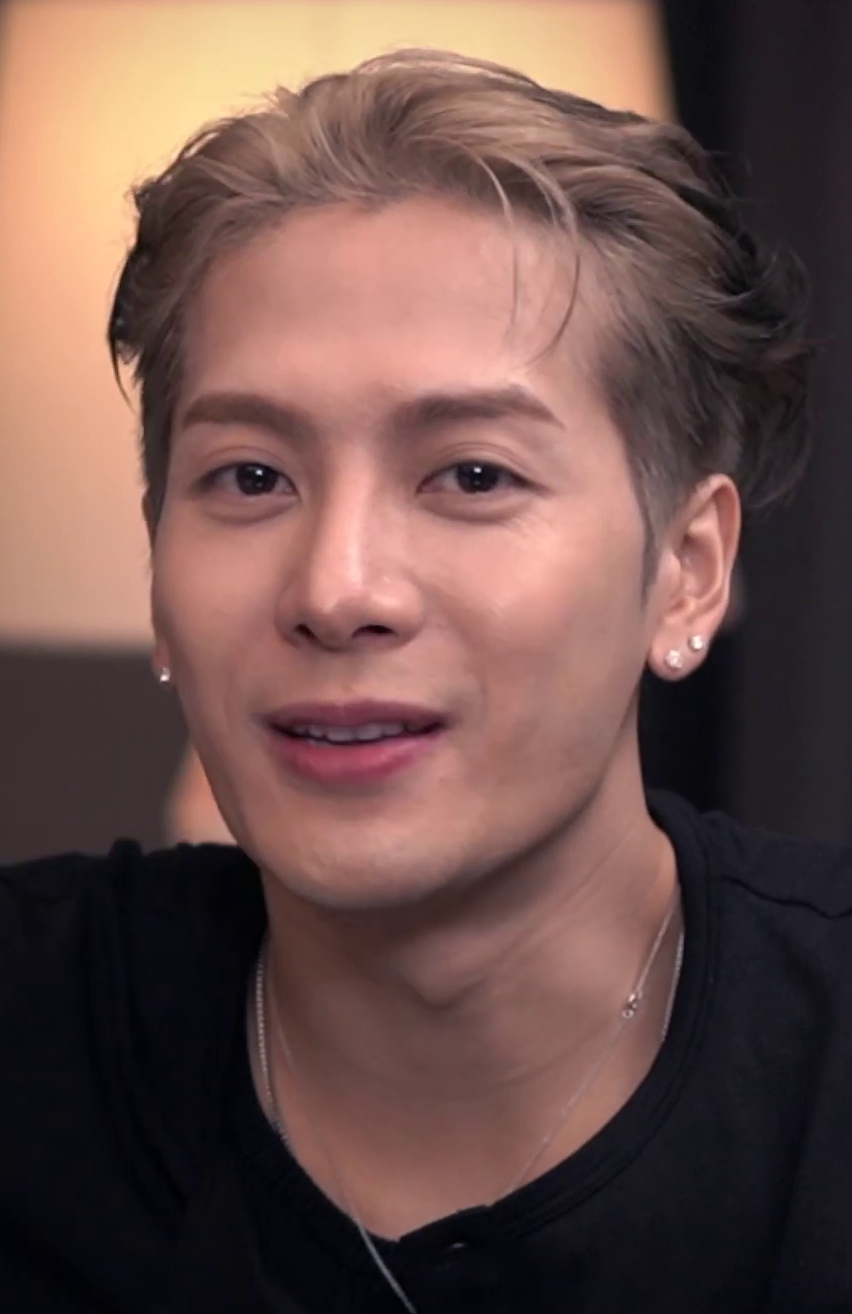 Close portrait of Jackson Wang mid-sentence in a black t-shirt and thin silver chain, hair dyed light blond and swept back