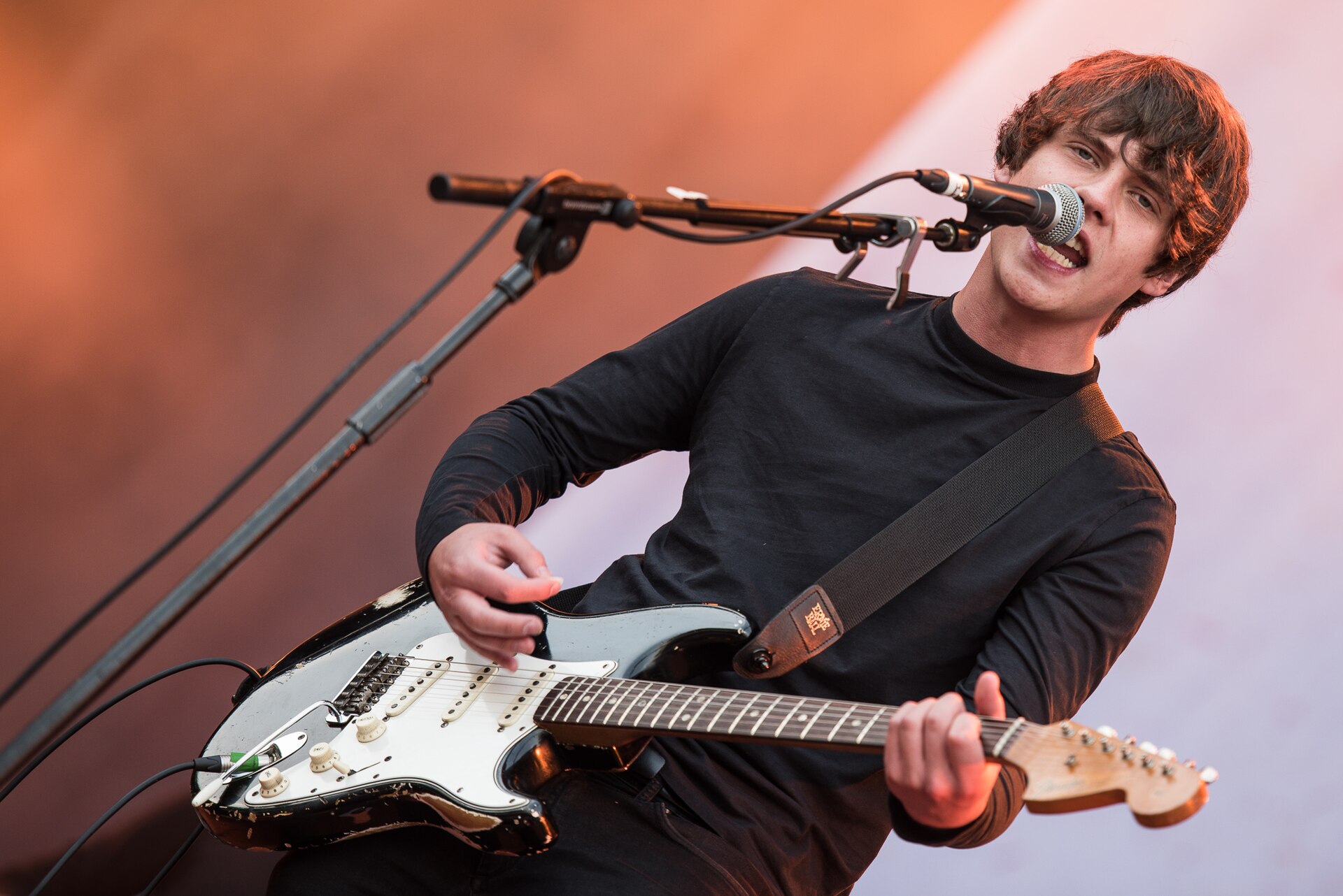Jake Bugg leans into a microphone mid-word with a battered black Stratocaster slung across a black long-sleeved top.