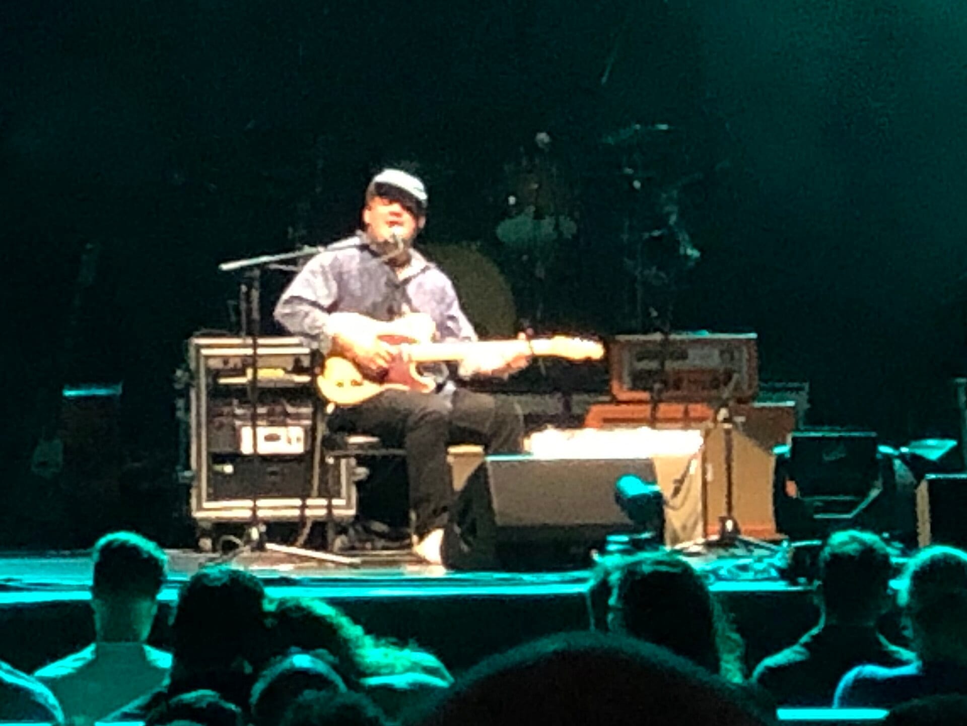 Jake Xerxes Fussell, wearing a light cap and button-up shirt, sits playing an electric guitar and singing into a microphone on a dim stage.
