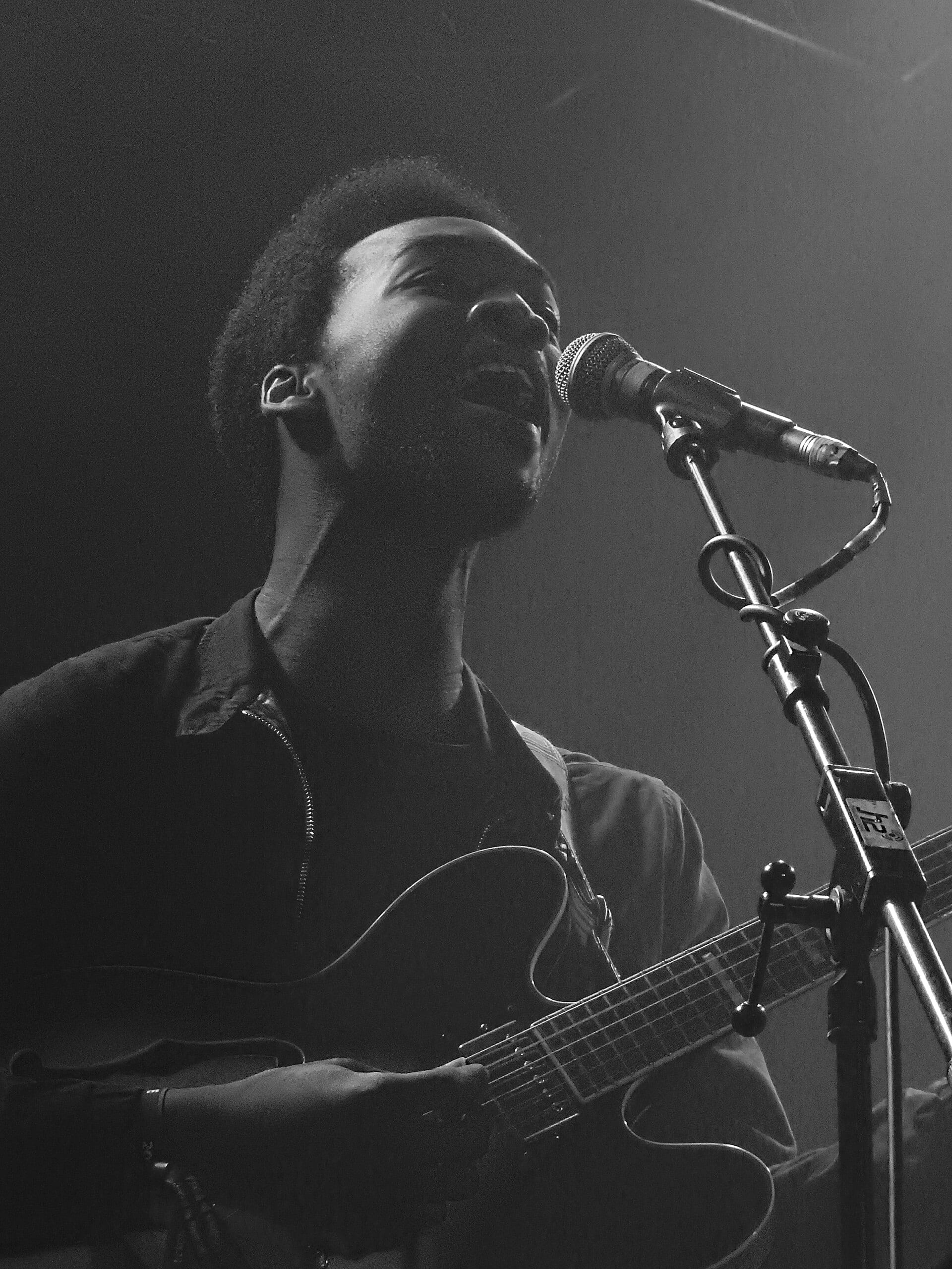 Jalen Ngonda sings into a microphone while playing a hollow-body electric guitar, lit dramatically from the side on a dark stage.