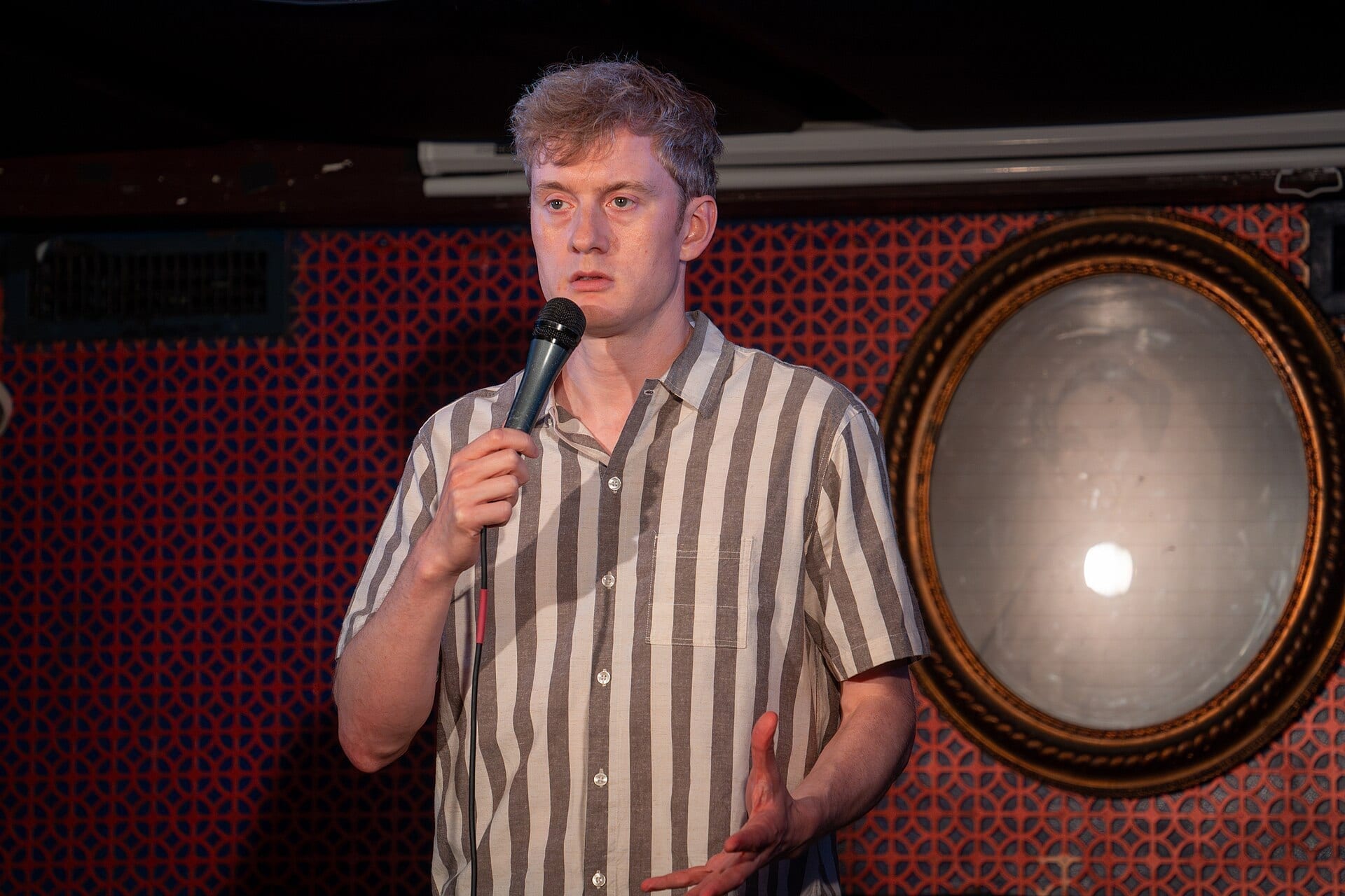 James Acaster holds a microphone on a small stage wearing a striped shirt, with a round mirror on the wall behind him.