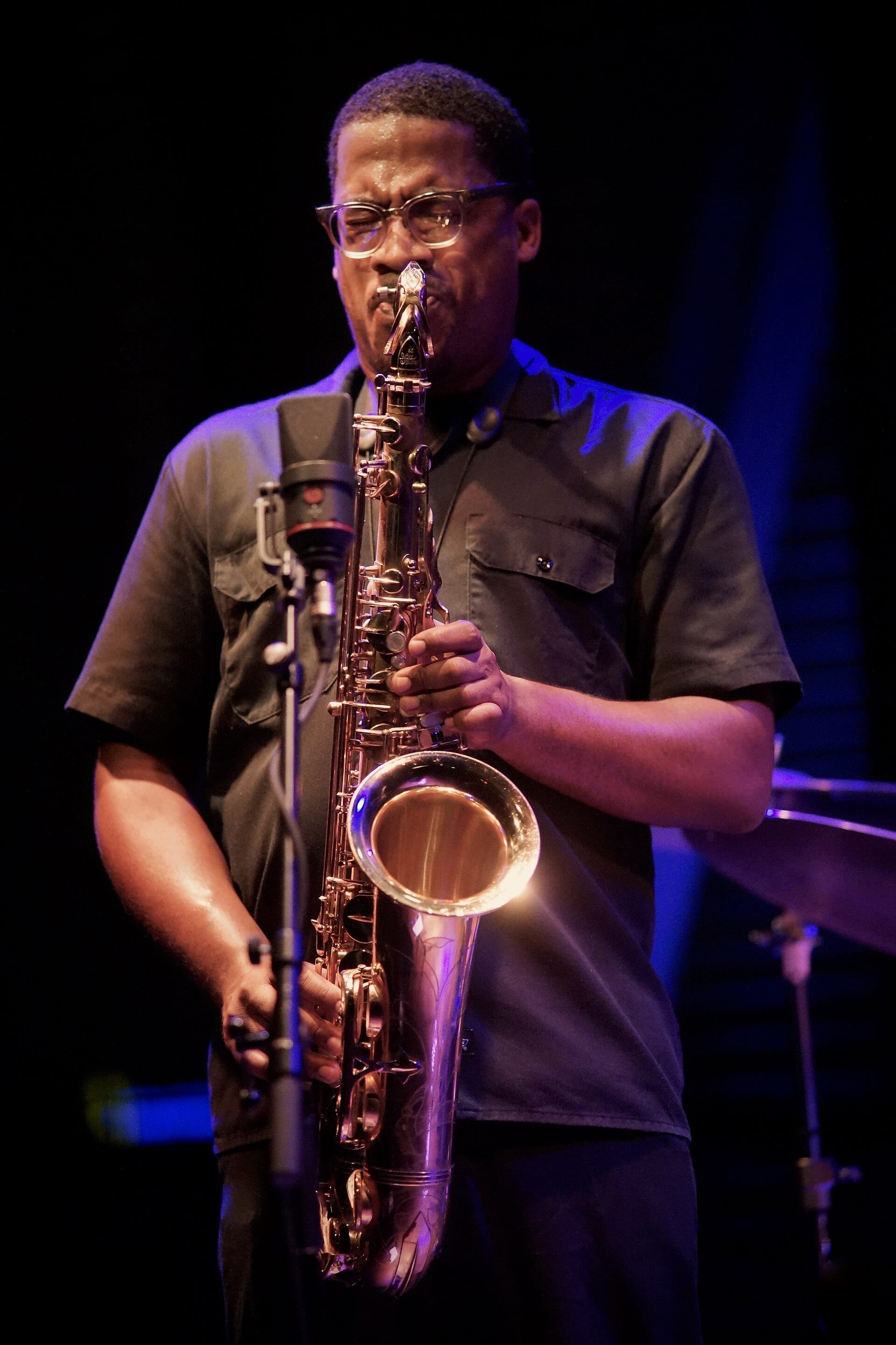 James Brandon Lewis plays a tenor saxophone under blue and amber stage light, wearing glasses and a dark work shirt.