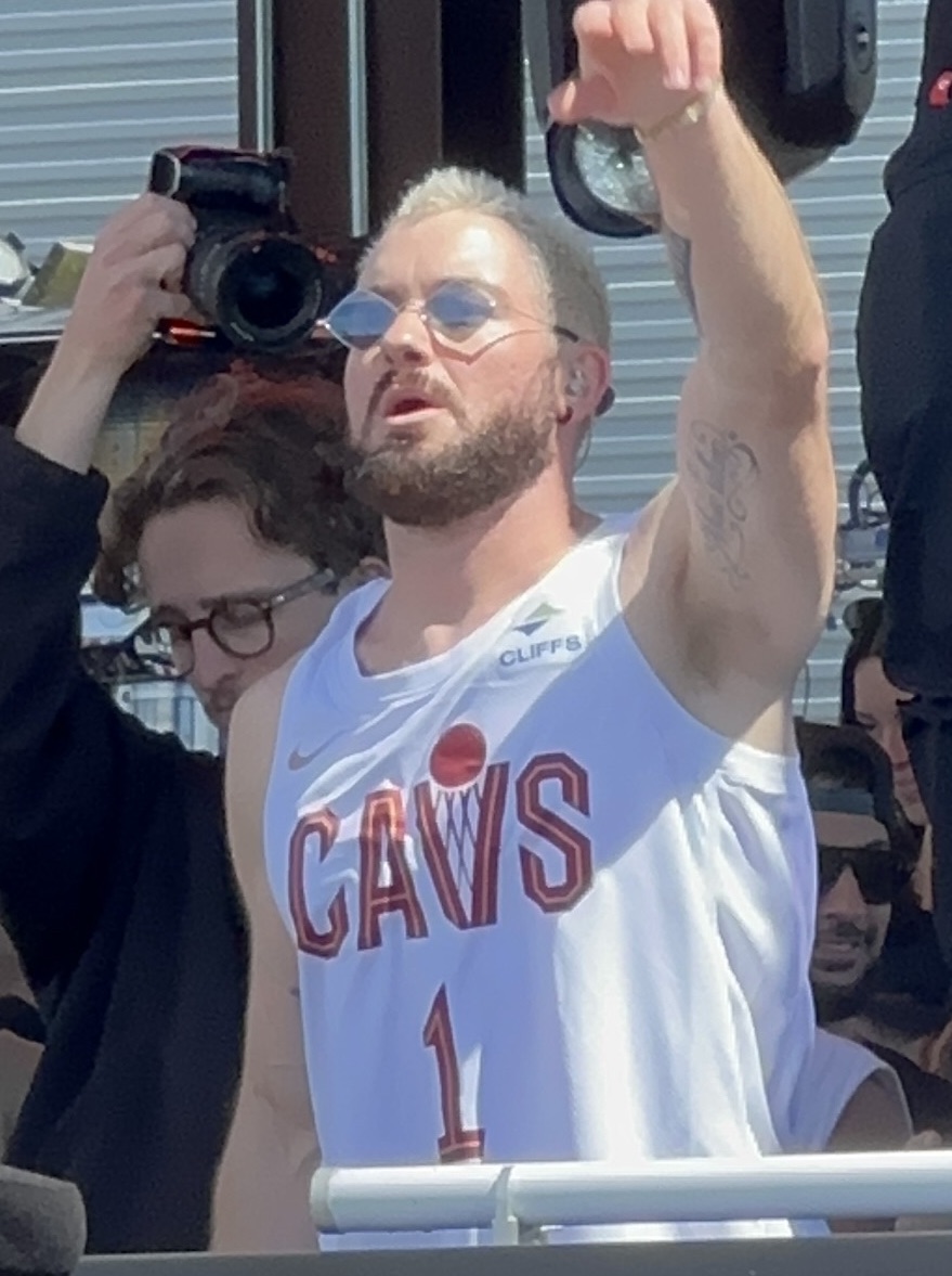 A DJ in blue-tinted round glasses and a white basketball jersey raises one arm behind the booth in daylight, a photographer shooting over his shoulder