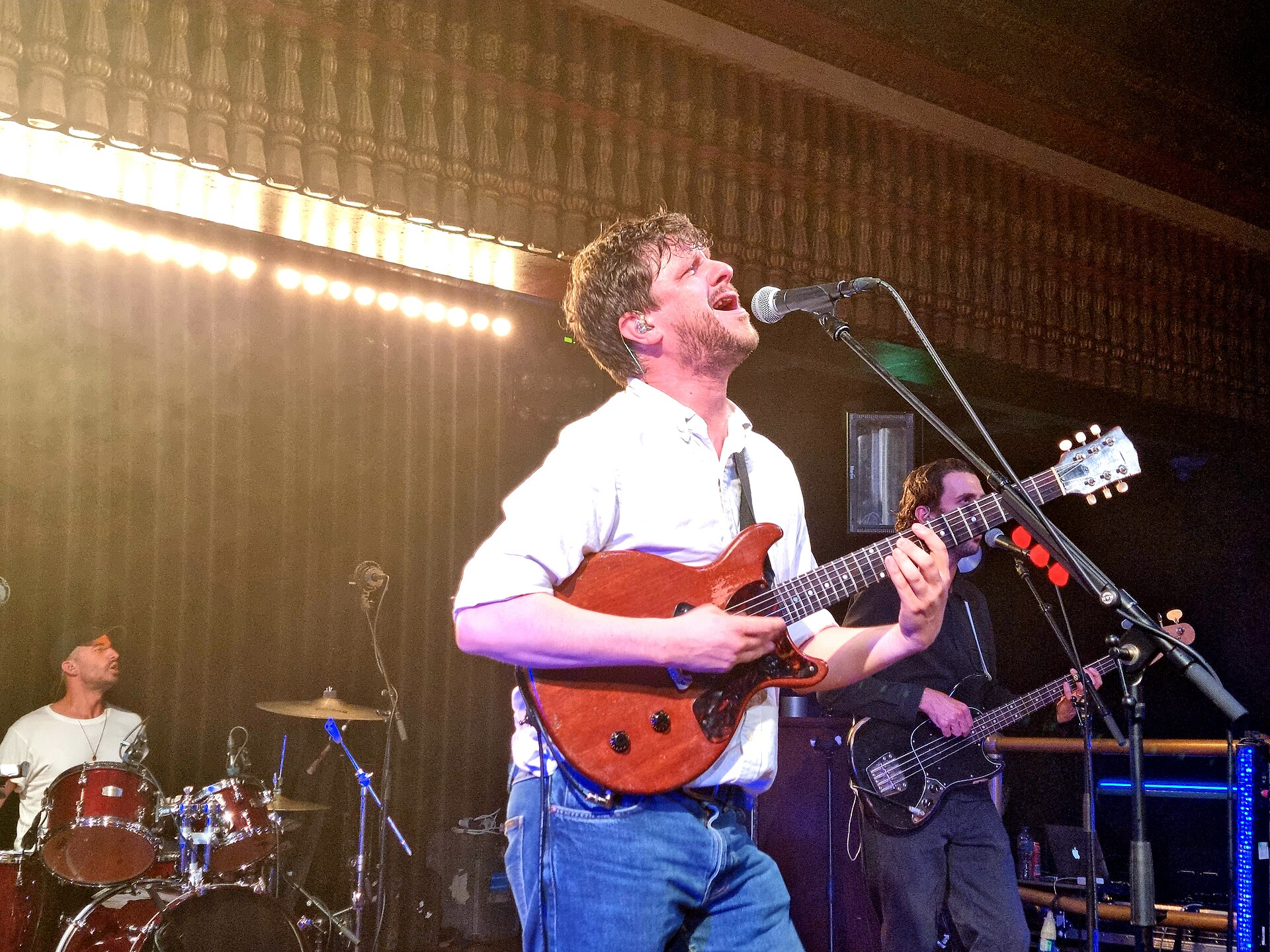 A singer in a white shirt and jeans plays a red electric guitar and sings up into a microphone, a drummer and bassist behind him on a small venue stage