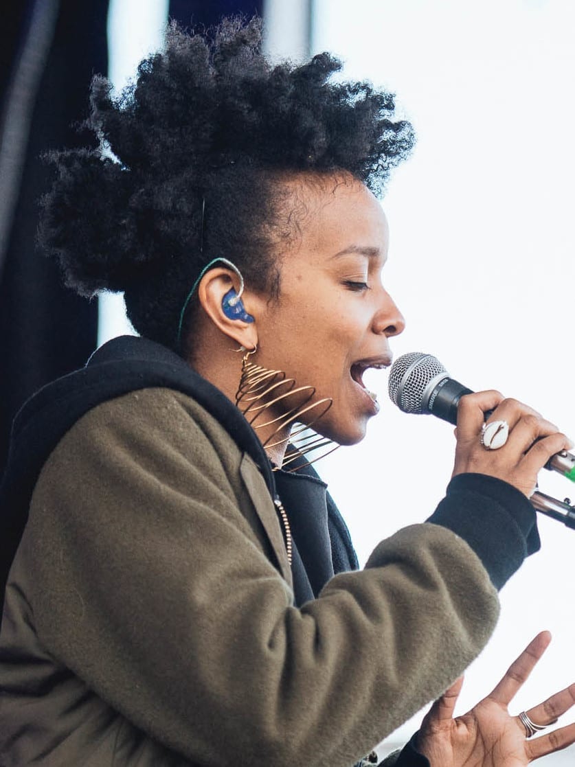 Jamila Woods, in profile with a curly updo and gold hoop earrings, sings into a microphone held close to her mouth.