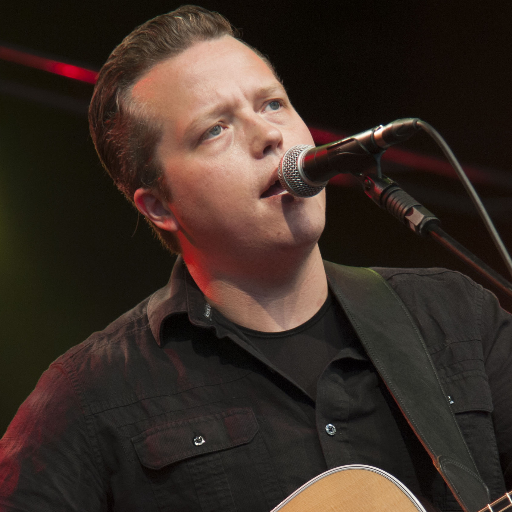 Jason Isbell sings into a microphone on a boom stand with an acoustic guitar strapped on, in a dark shirt under red stage light.