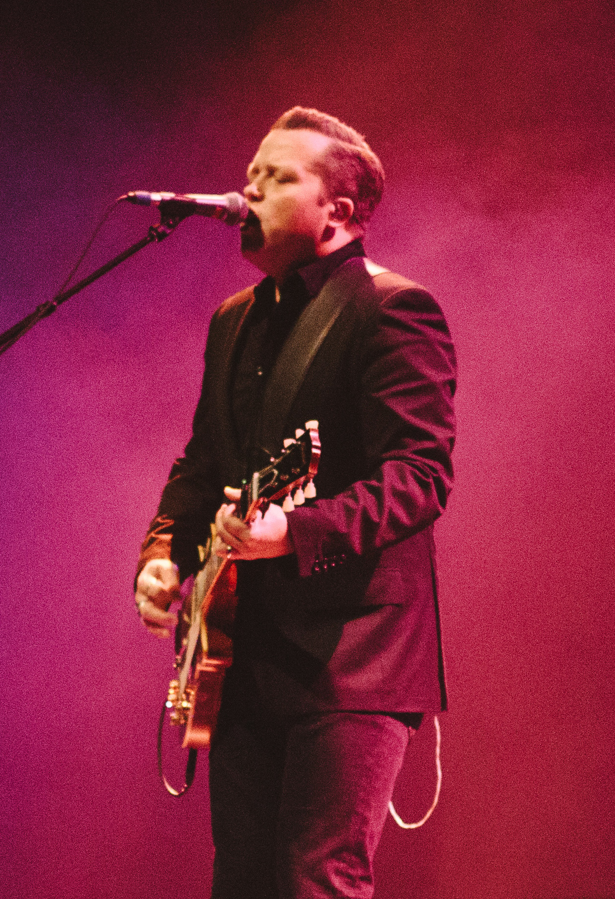 Jason Isbell sings into a mic on a boom arm with a red hollow-body guitar, in a dark suit jacket against a deep pink backdrop.