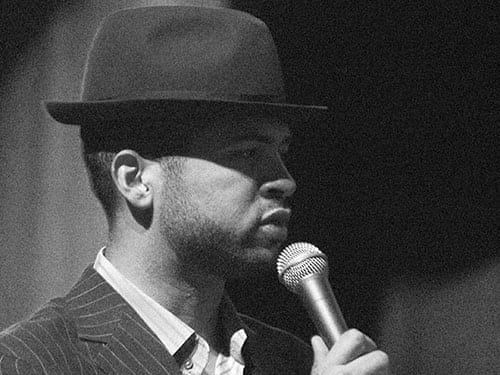 A black-and-white photo of Jason Moran, wearing a fedora and pinstriped jacket, speaking into a microphone in profile.