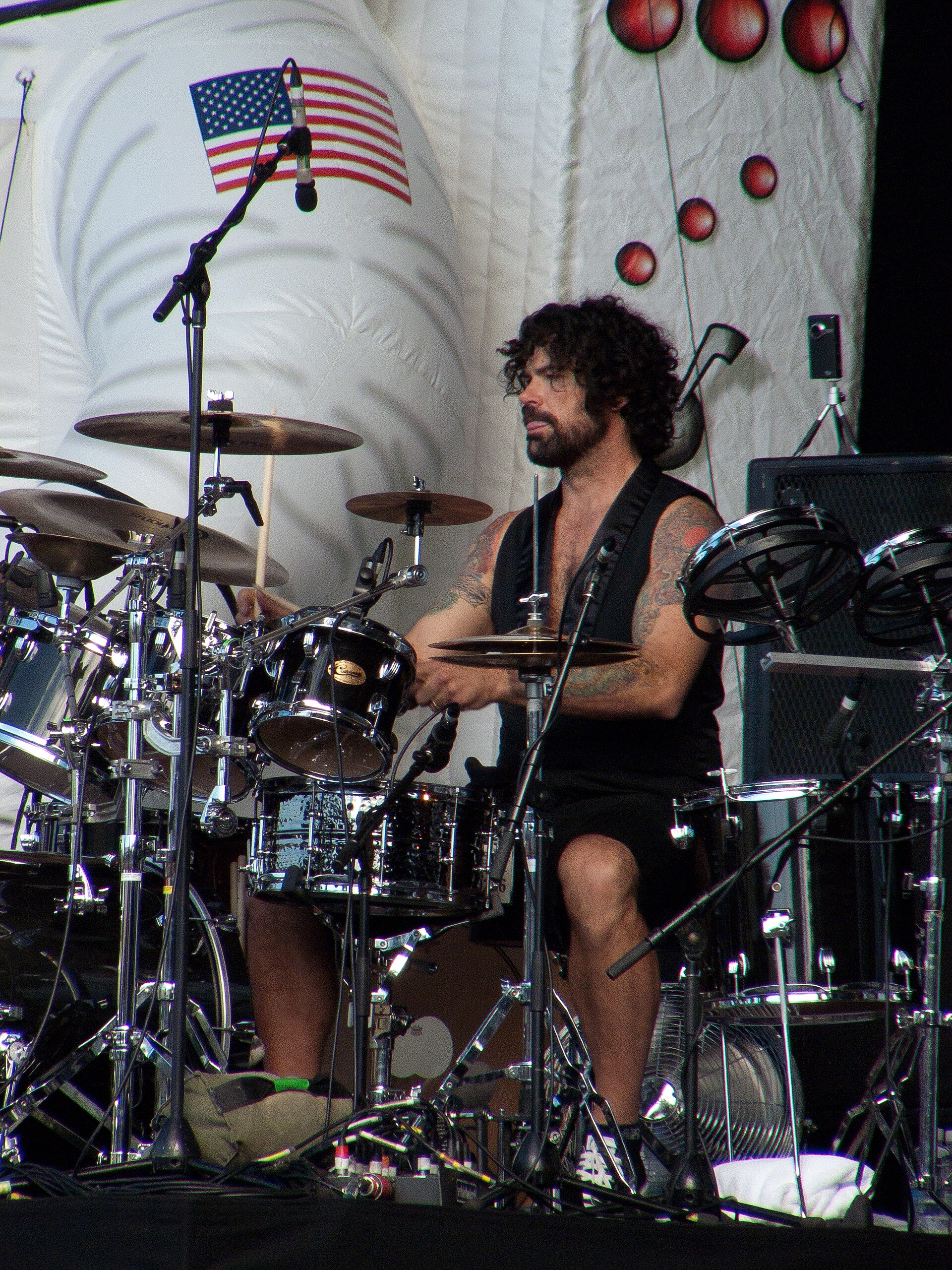 Jay Lane plays a large drum kit in a black vest and shorts, curly hair and beard, a US flag pinned to the white backdrop behind him.