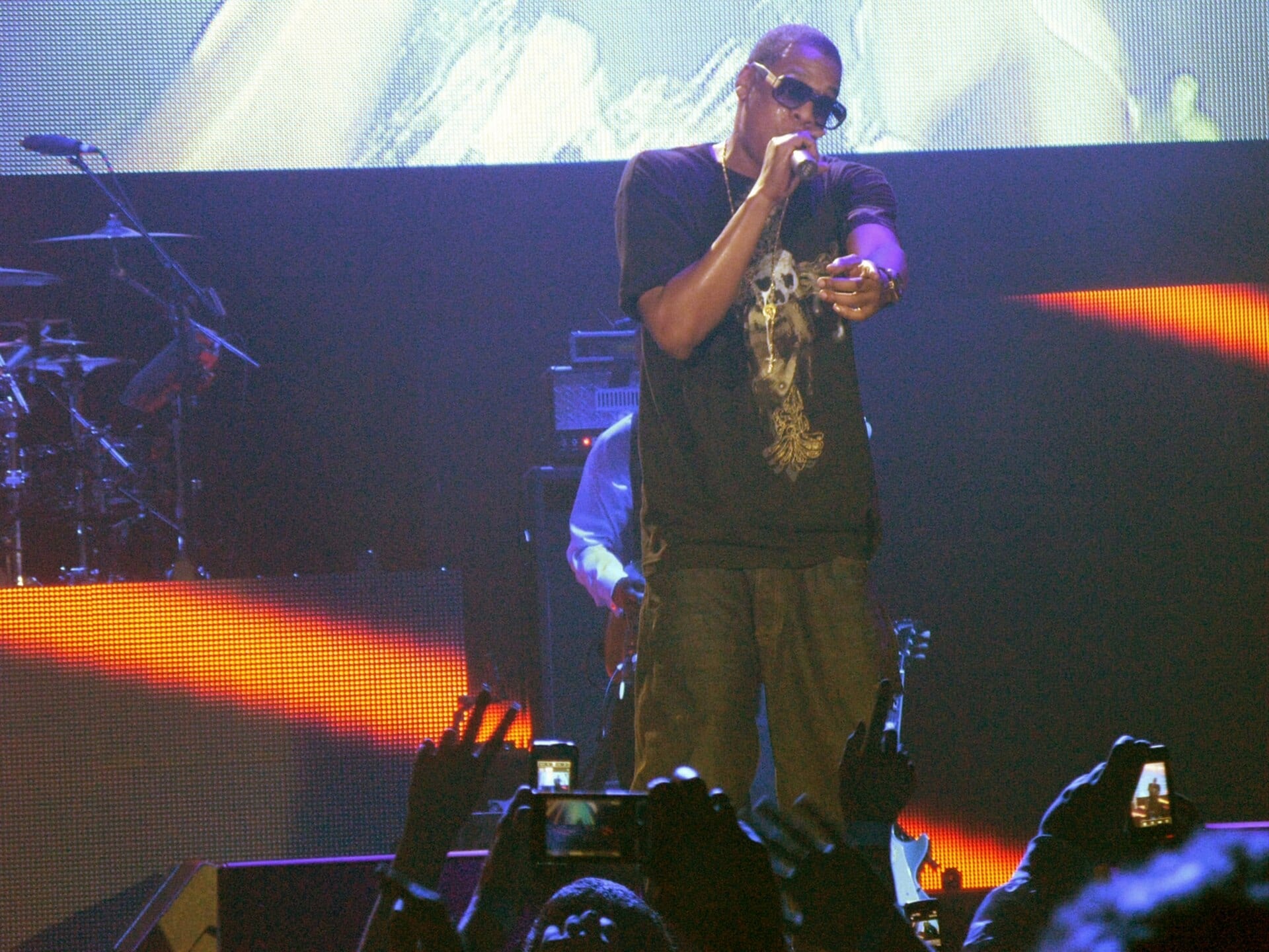 Jay-Z performing on stage, wearing sunglasses and a dark graphic T-shirt, holding a microphone and gesturing toward the crowd, with fans holding up phones in the foreground.