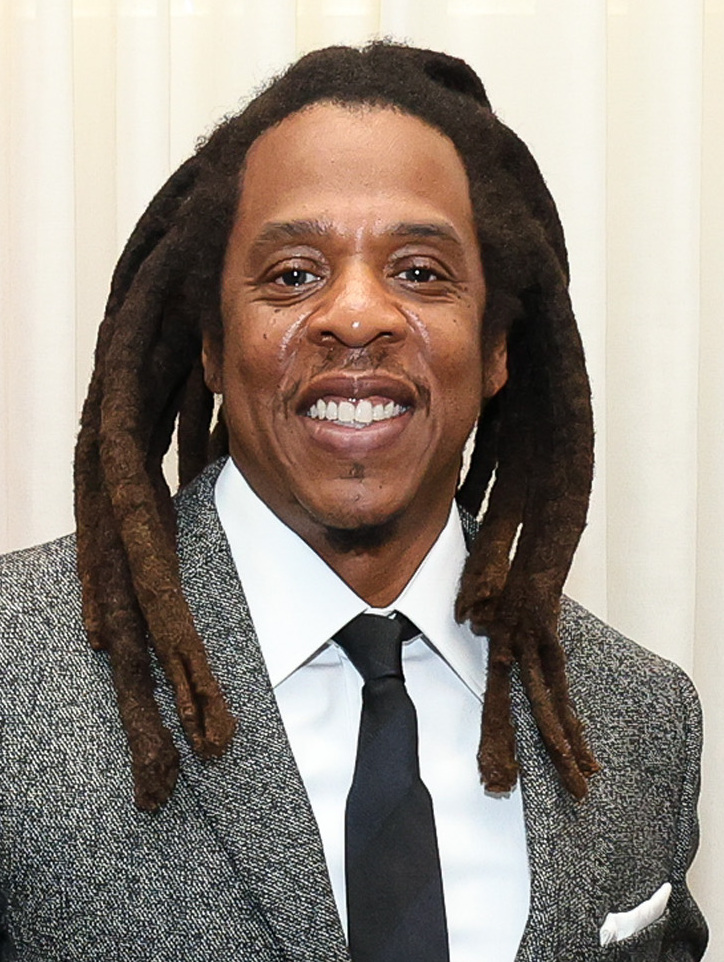 Jay-Z smiles for the camera in a grey tweed jacket, white shirt and dark tie, locs falling past his shoulders.