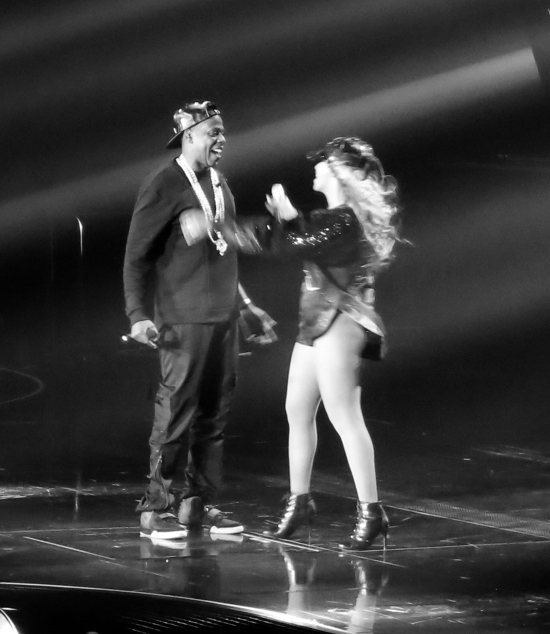 Jay-Z and Beyonce share a moment on stage in this black-and-white photo, Jay-Z in a cap and chains, Beyonce mid-dance in a sequined outfit.