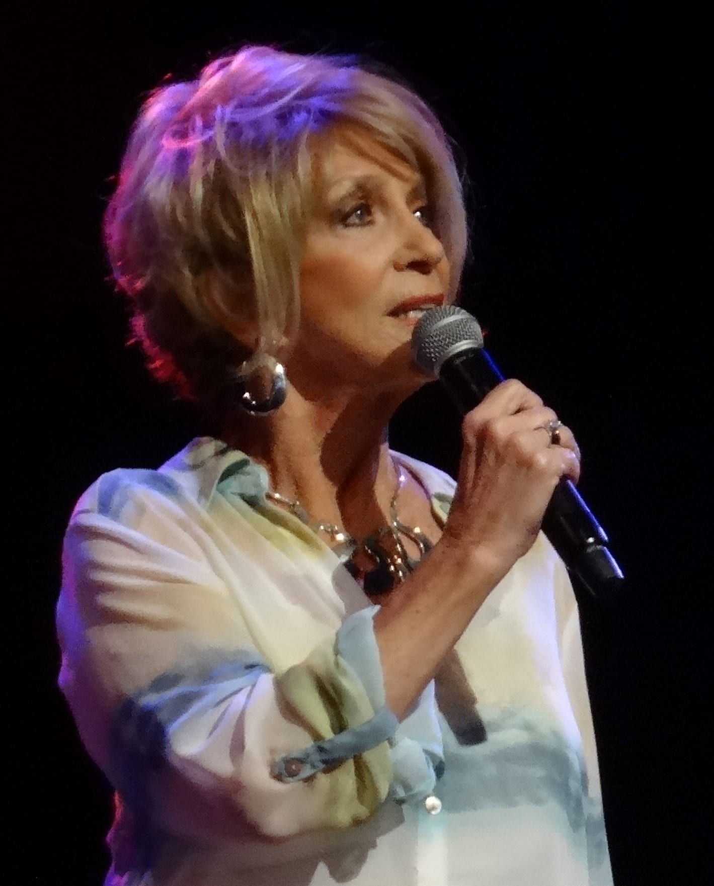Jeannie Seely, in a flowing patterned blouse and statement earrings, sings into a handheld microphone against a dark backdrop.