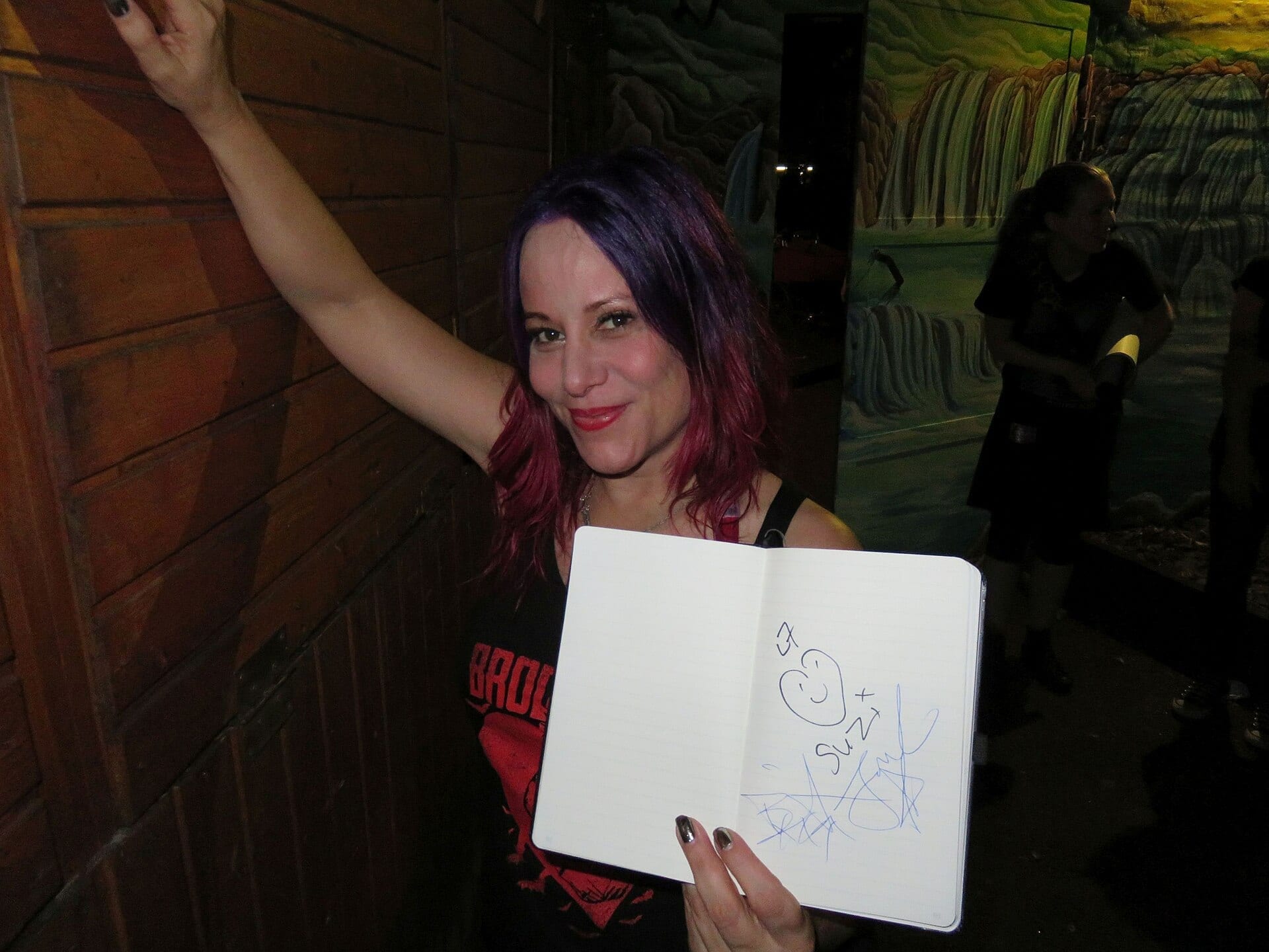 Jennifer Finch, with purple and red streaked hair, smiles and holds up a signed notebook after a show.