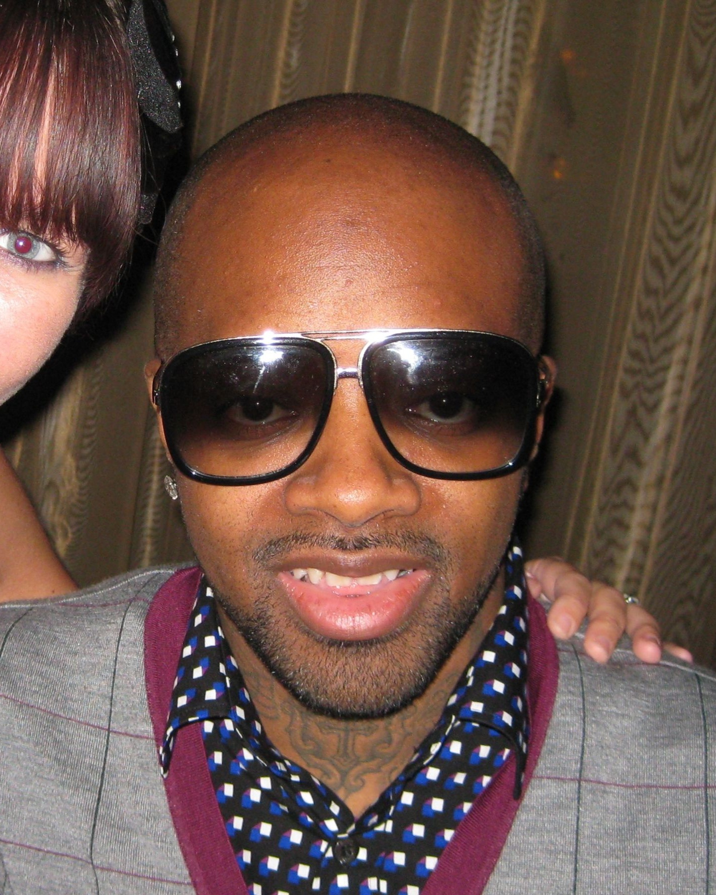 Jermaine Dupri stands at a conference in dark sunglasses and a black jacket over a patterned shirt, hands clasped in front of him.