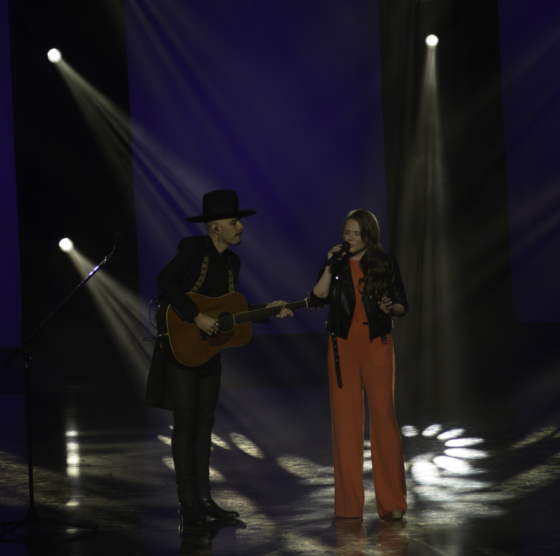 Jesse and Joy Huerta perform as a pair on a dark stage, he in a black wide-brimmed hat playing an acoustic guitar, she singing into a handheld microphone in an orange jumpsuit and leather jacket.