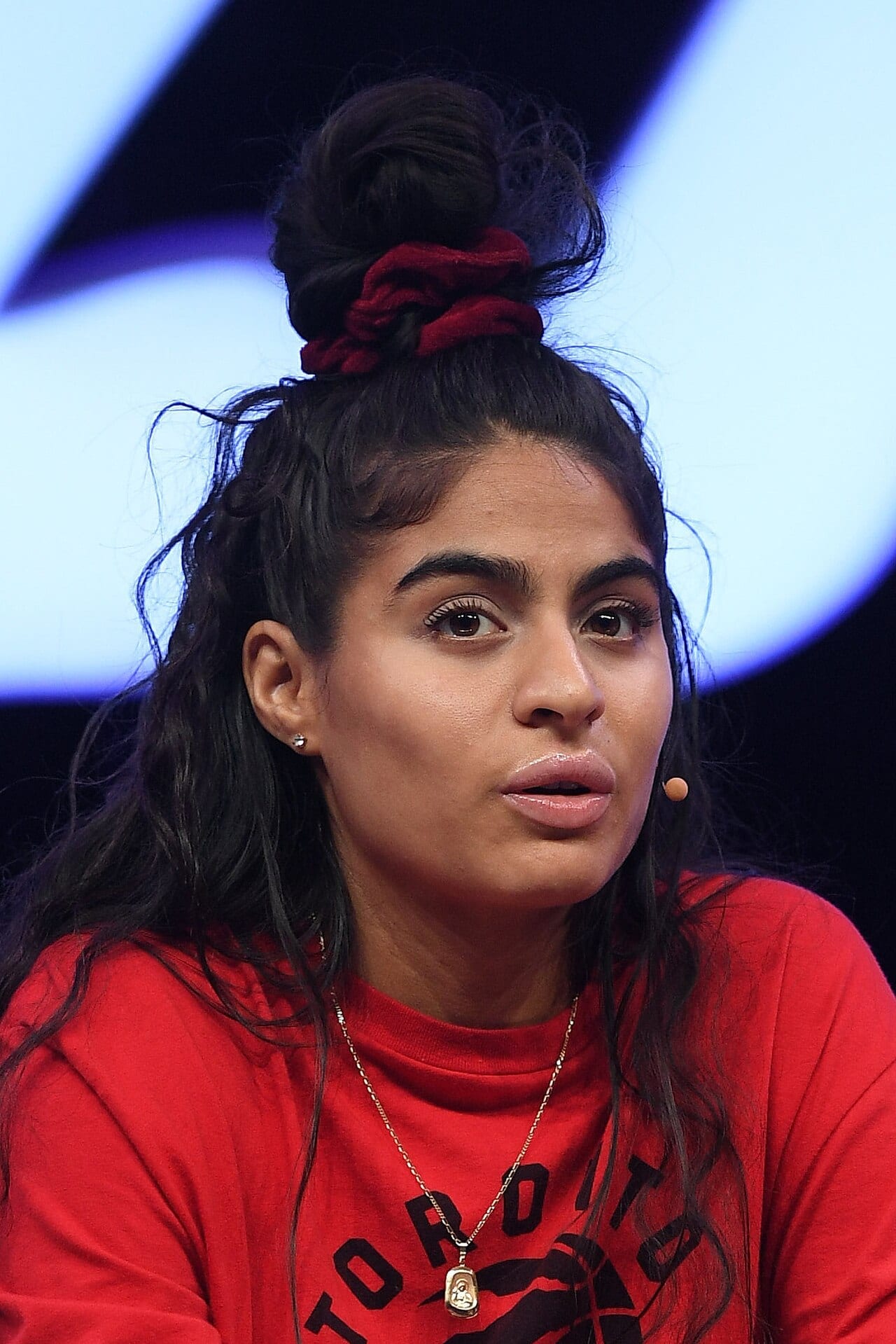 Jessie Reyez, in a red Toronto Raptors shirt with her hair in a top knot, speaks while wearing a stage earpiece.