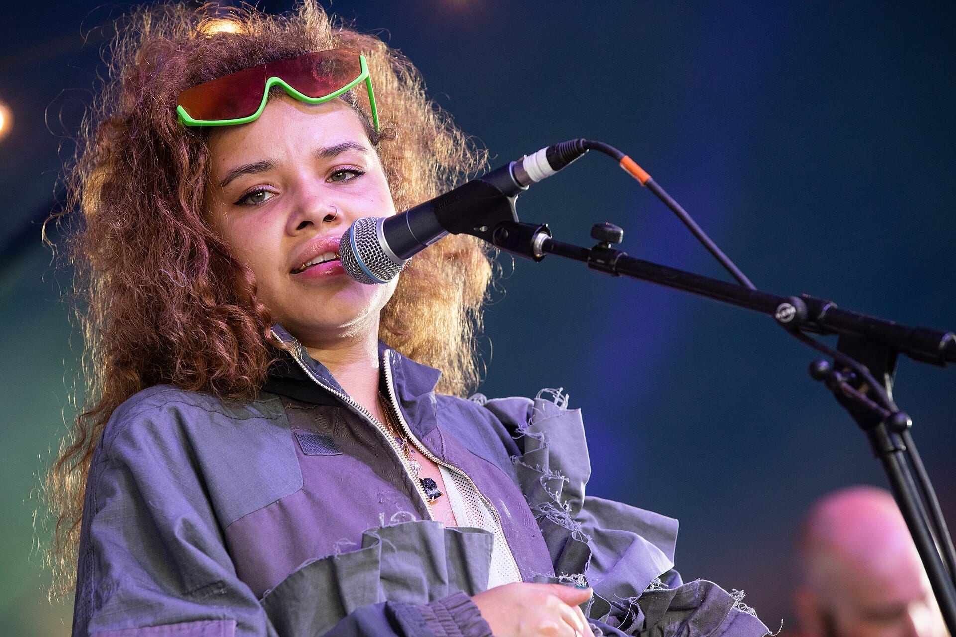 JGrrey sings close to a microphone, curly hair loose, sunglasses pushed up onto her head.