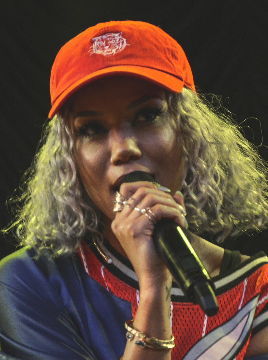 A singer with blonde curly hair and an orange cap sings into a handheld microphone, close in frame against a dark background