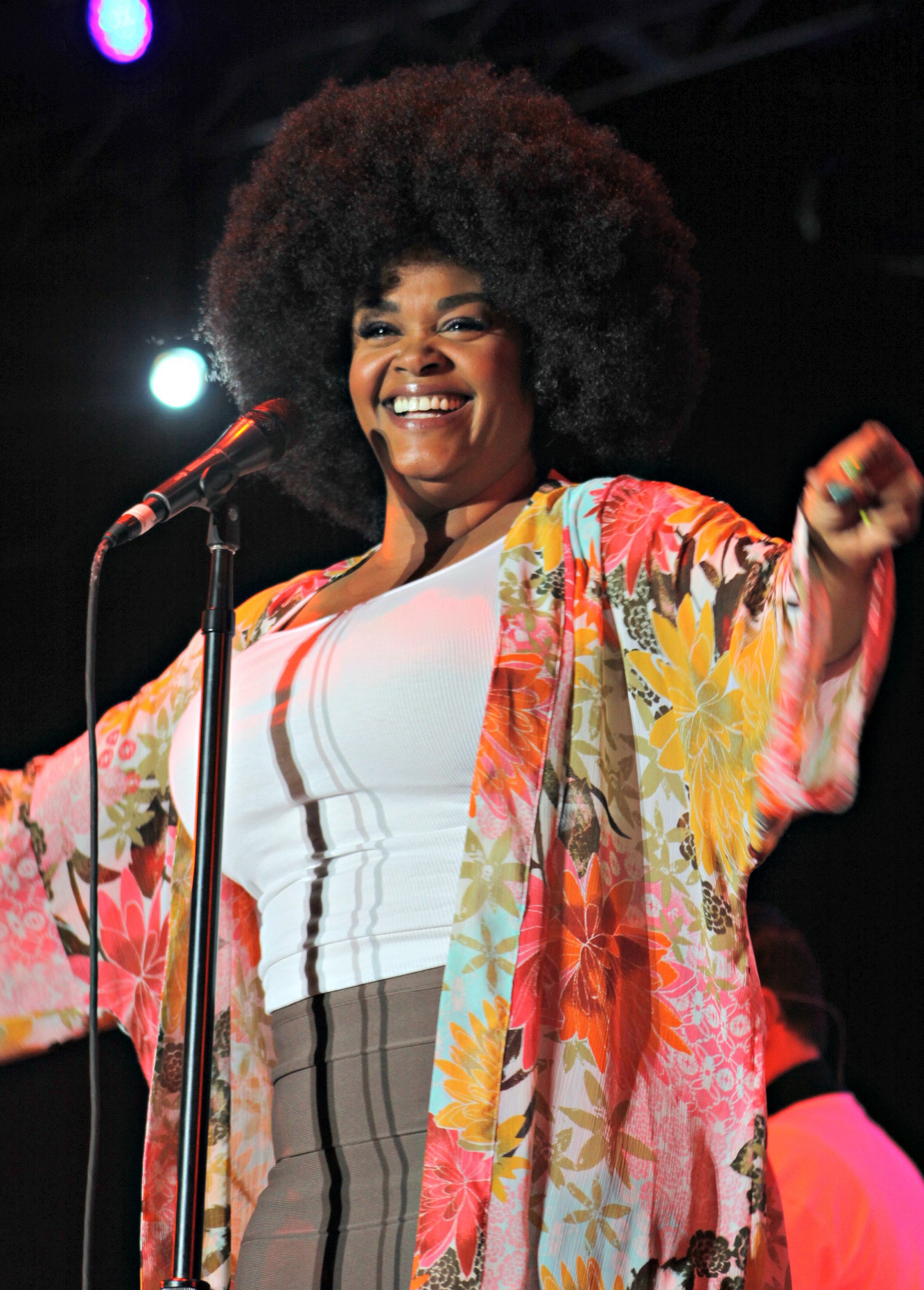 Jill Scott laughs behind a microphone stand with both arms spread, wearing a floral kimono over a white top, her hair worn in a large afro.