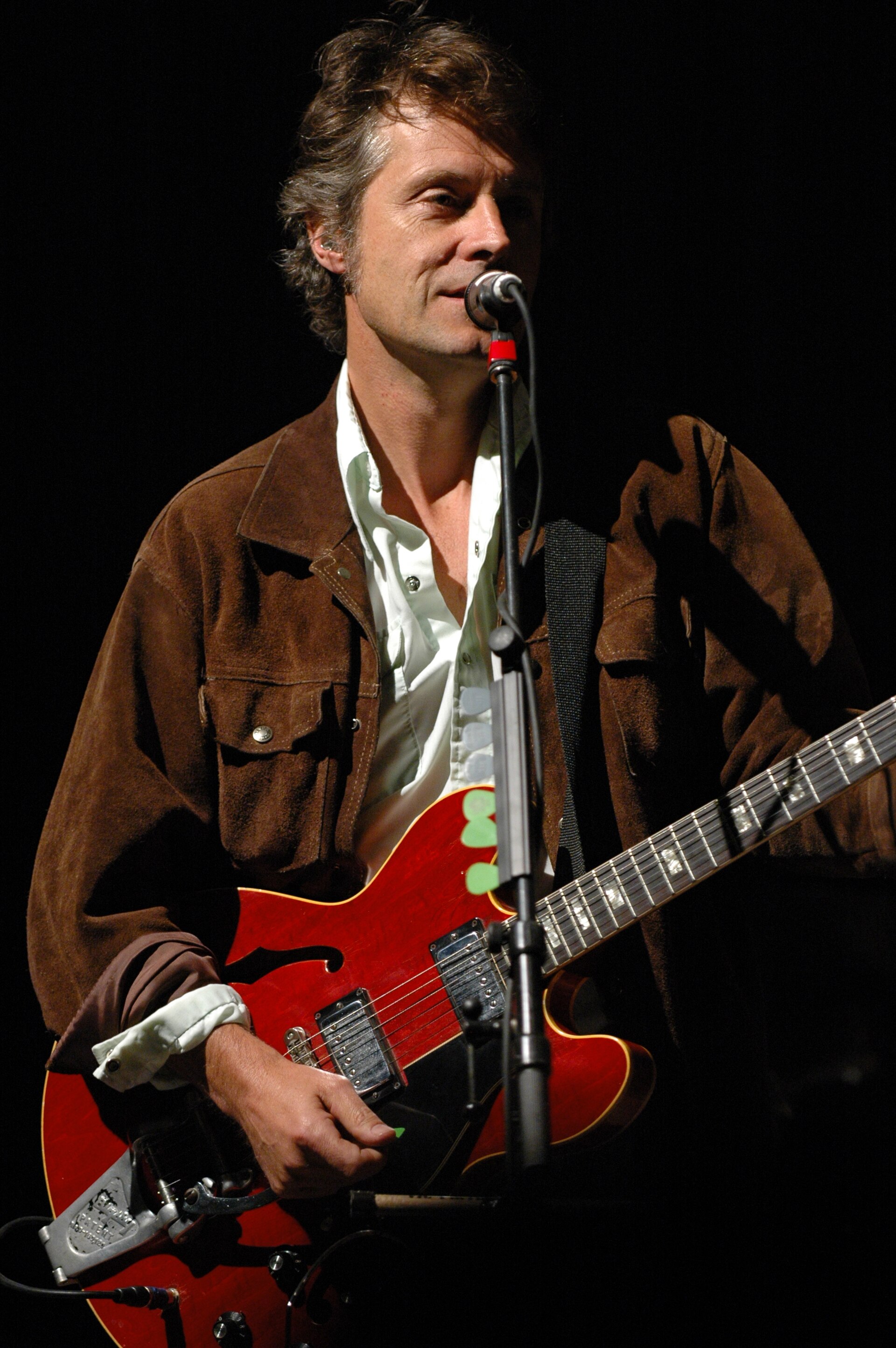 Jim Cuddy at a microphone in a brown suede jacket over a pale shirt, playing a red hollow-body electric guitar against a black backdrop