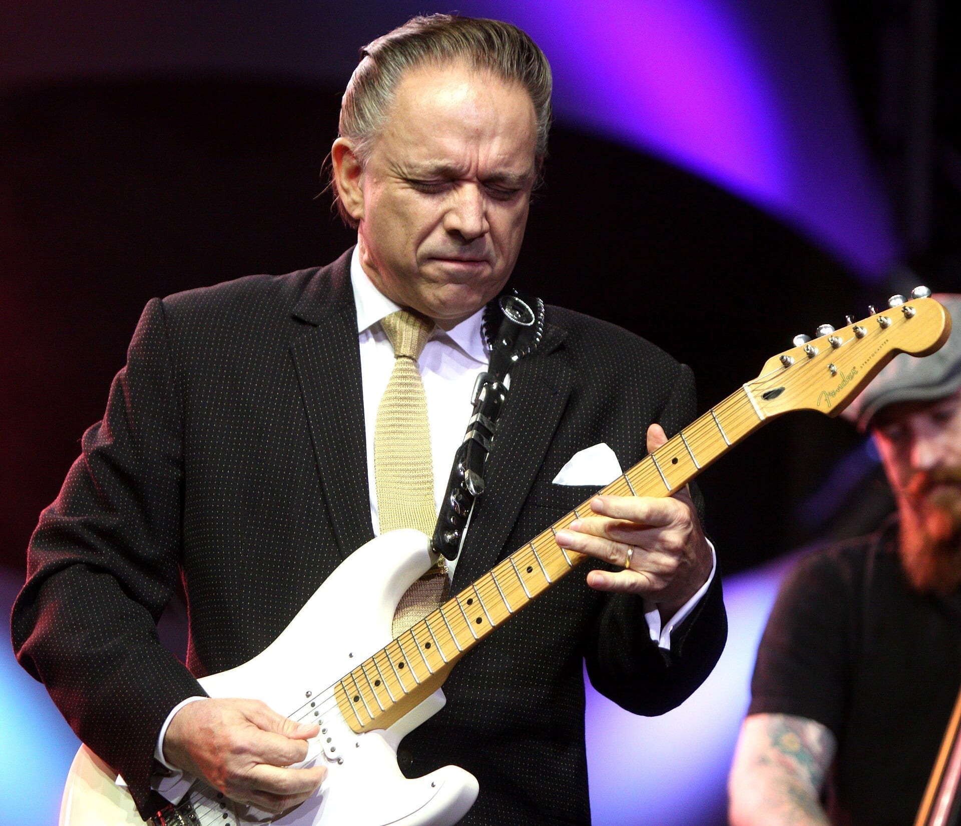 Jimmie Vaughan plays a white Fender Stratocaster on stage in a pinstriped suit and tie, eyes closed in concentration, with a bandmate visible behind him.