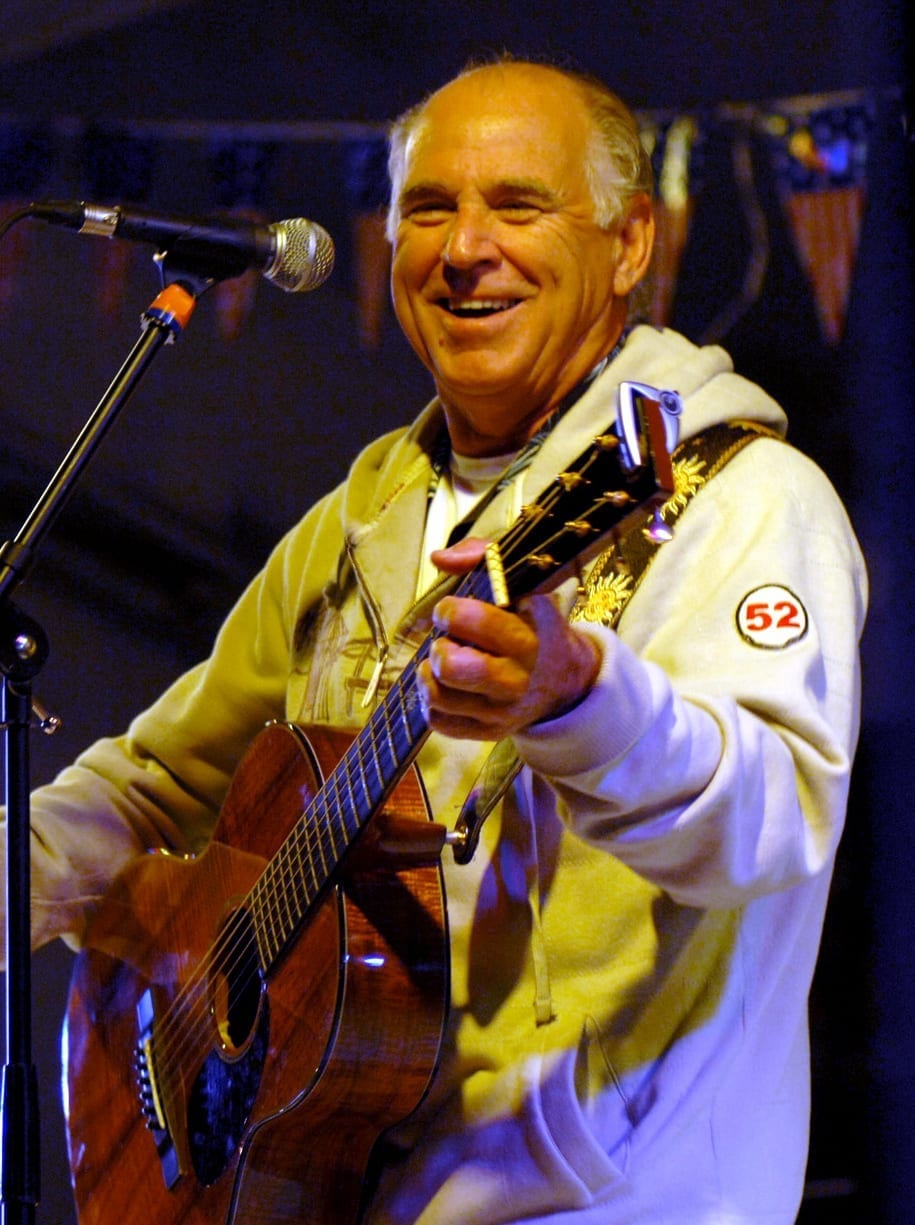 Jimmy Buffett smiles while playing an acoustic guitar into a microphone on stage, wearing a cream hoodie with a numbered patch on the sleeve.