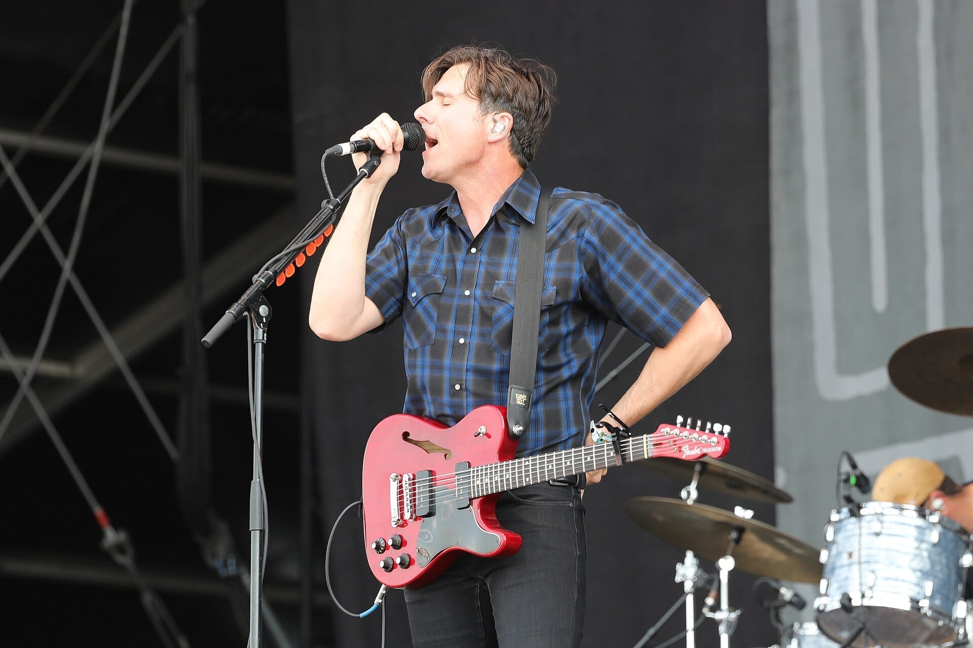 A Jimmy Eat World band member sings into a microphone while holding a red electric guitar, with a drummer visible behind him on an outdoor festival stage.