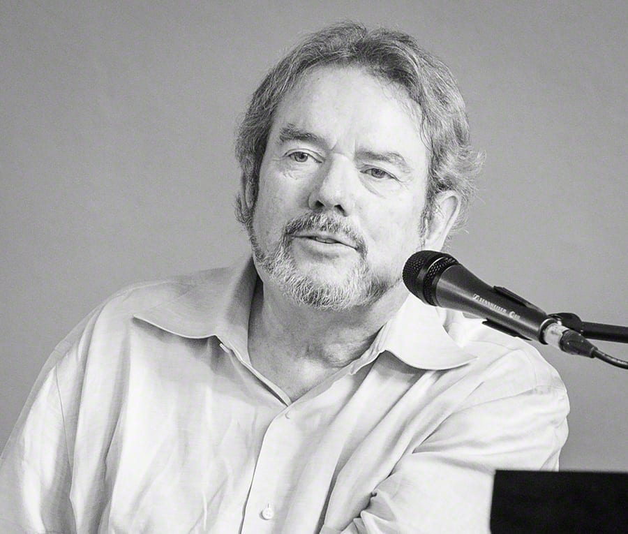 A black and white portrait of songwriter Jimmy Webb speaking near a microphone, wearing a light collared shirt.