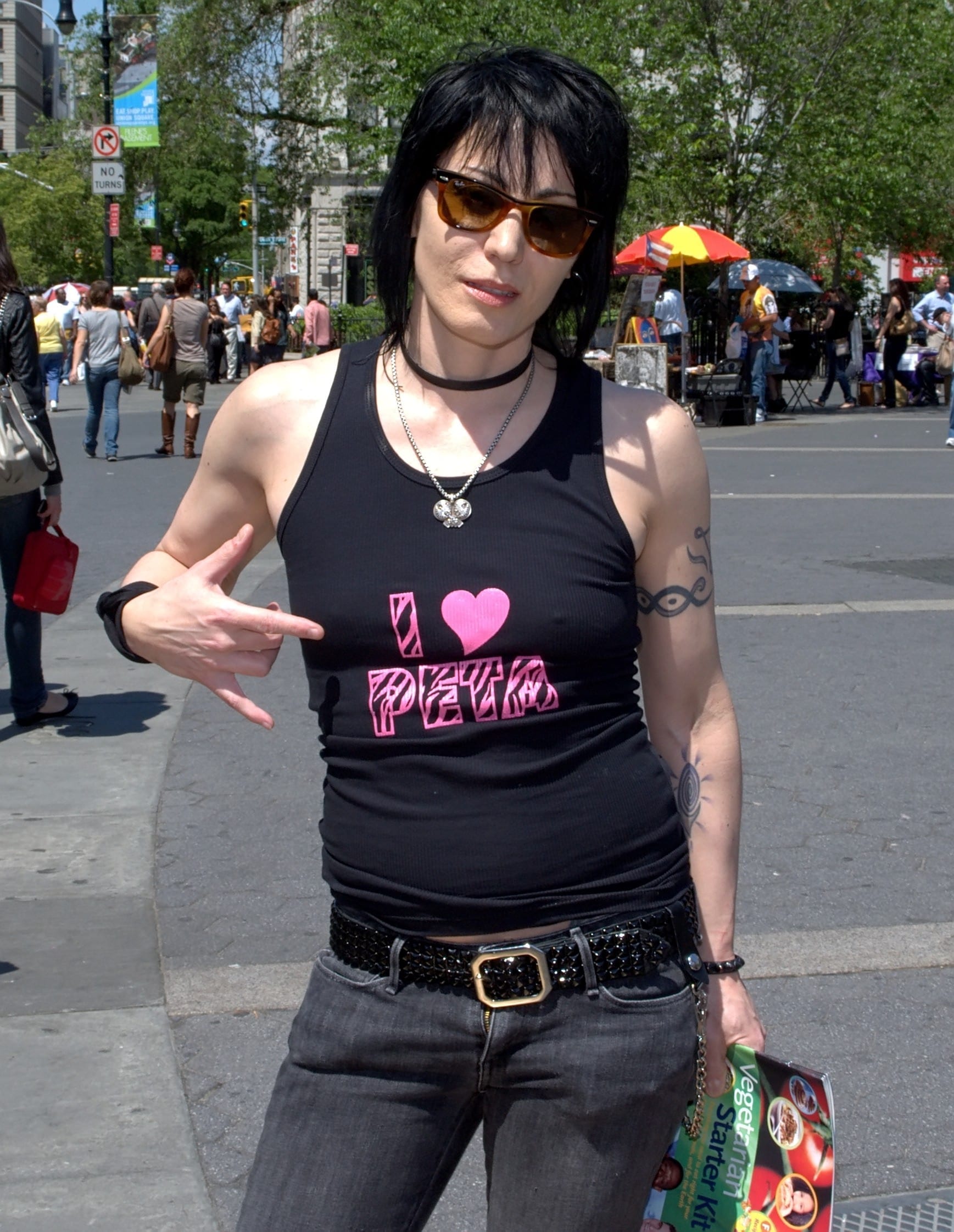 Joan Jett stands on a city street in sunglasses and a black tank top reading I Love PETA, pointing at the shirt with visible arm tattoos, dark hair falling across her face.