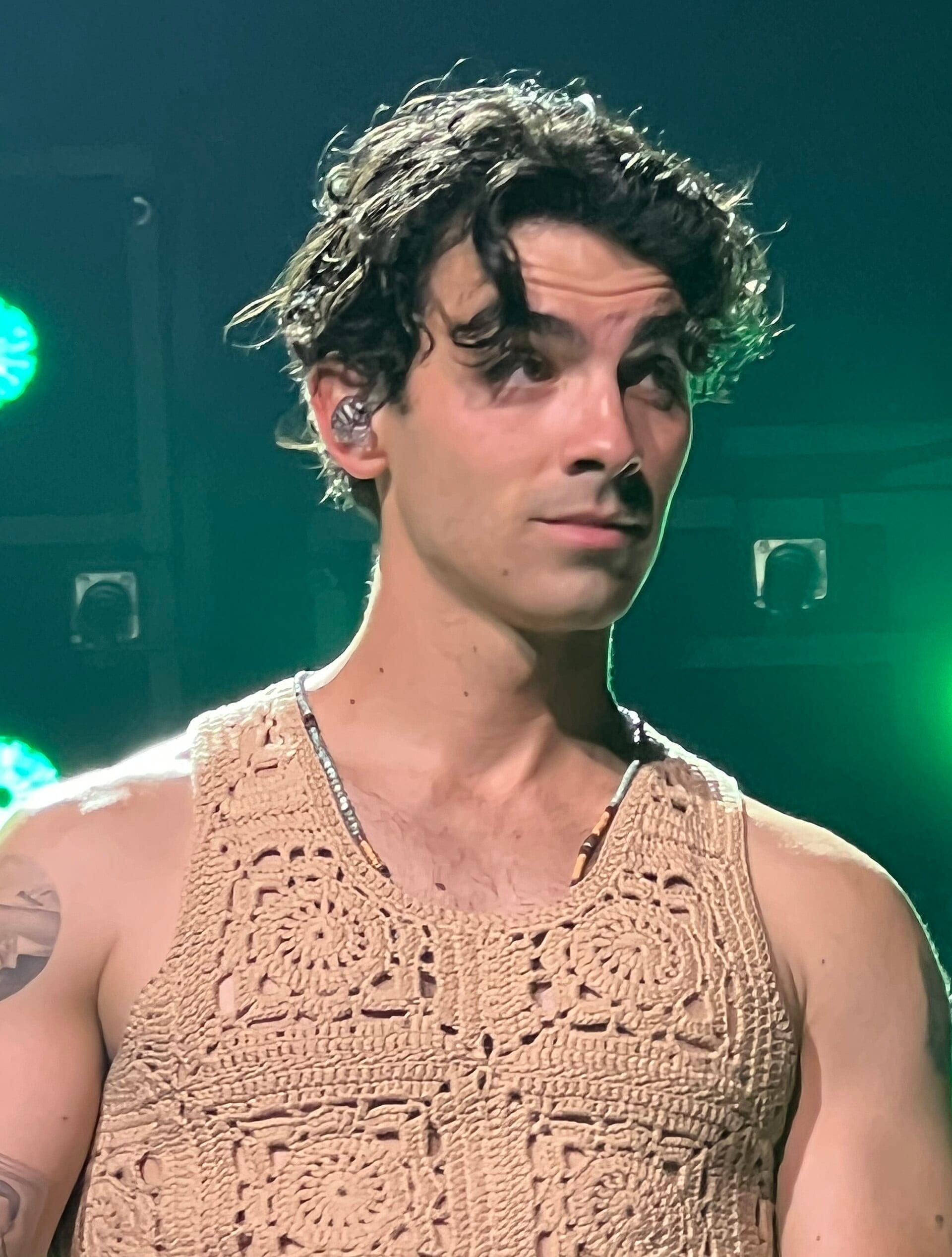 A close-up of Joe Jonas on stage, his curly dark hair falling over one eye, wearing a tan crocheted tank top, lit by green stage light.