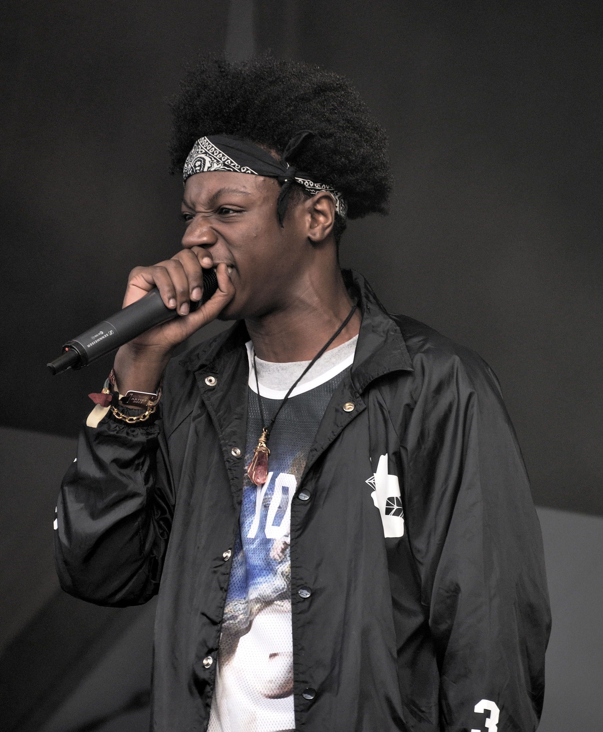 Joey Bada$$, wearing a bandana over his afro and a black jacket, sings into a handheld microphone on stage.