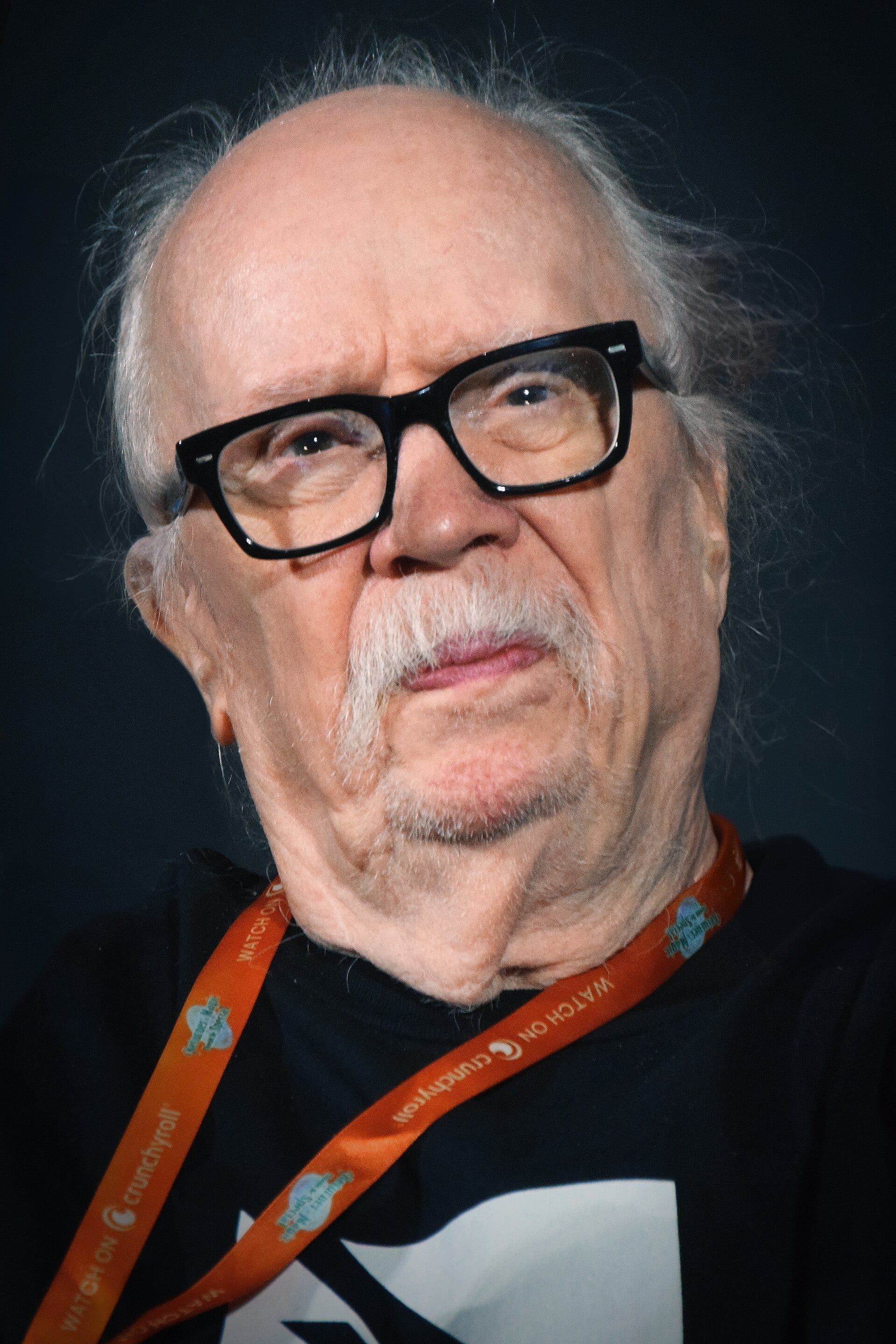 John Carpenter, wearing thick black glasses and a gray mustache, looks toward the camera in a close-up portrait with an orange lanyard around his neck.