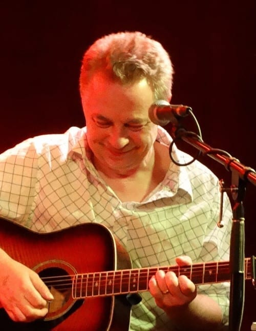 John Douglas of the Trashcan Sinatras smiles down at his acoustic guitar while singing into a microphone under red stage light.