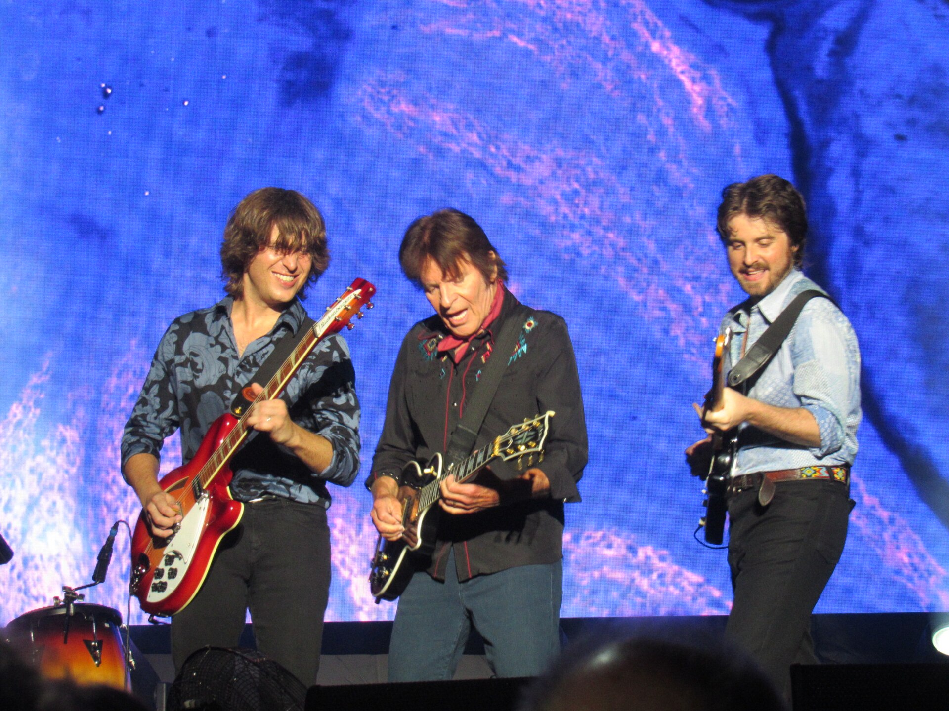 John Fogerty plays guitar centre-stage between two younger guitarists, all three grinning, against a blue and pink projected backdrop.