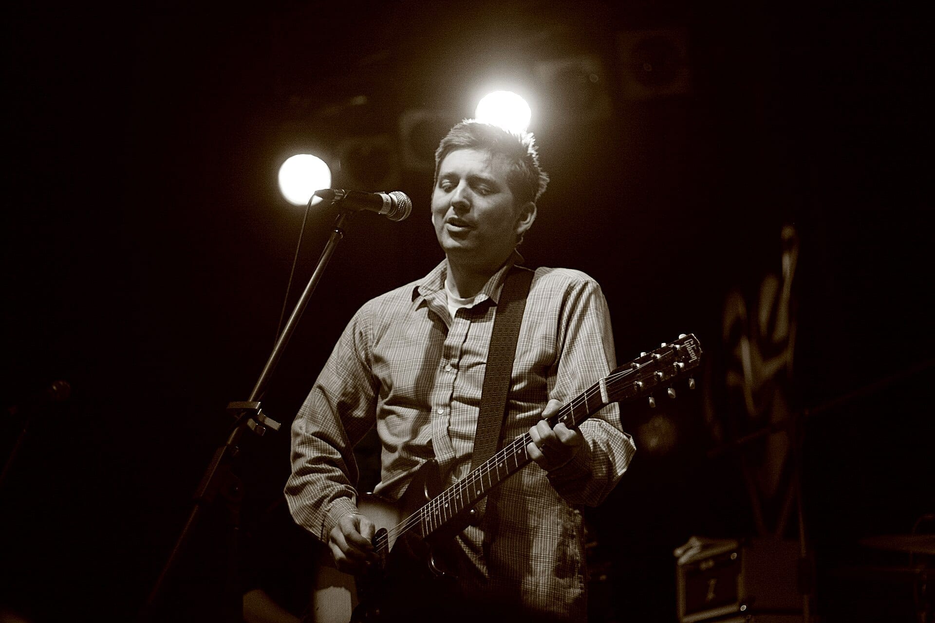 John K. Samson sings into a microphone while playing an electric guitar on a dim stage, lit from above in a sepia-toned photograph.