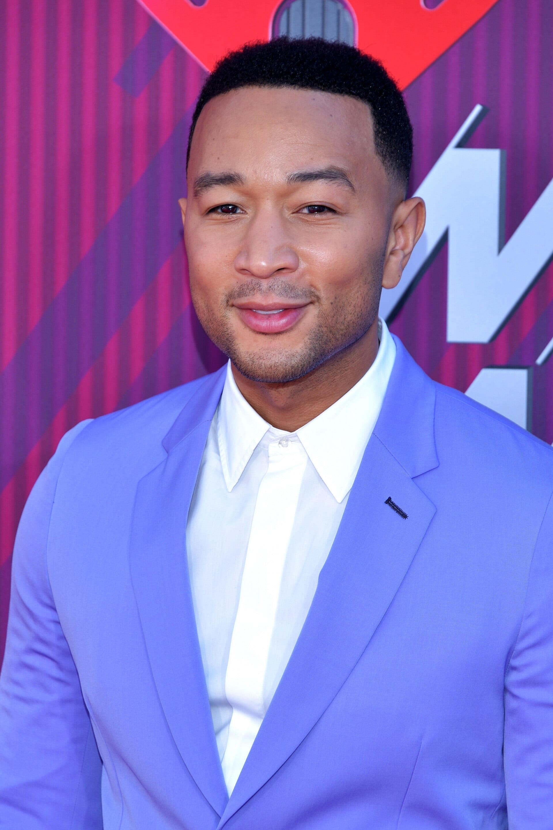 John Legend smiles softly in a close portrait, wearing a periwinkle-blue suit jacket over a white collared shirt.
