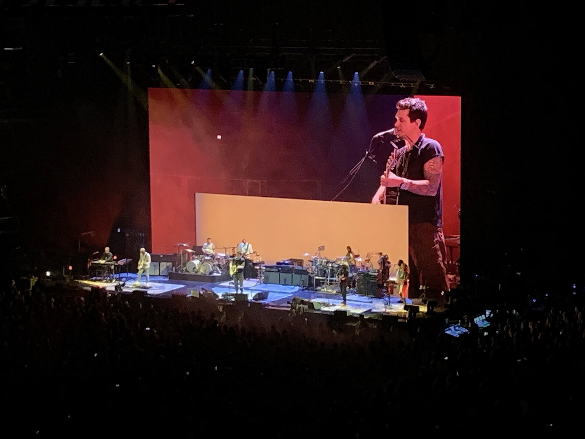 A view from the back of an arena floor as John Mayer plays guitar alone at the front of a wide stage, his band behind him and a large screen alongside.
