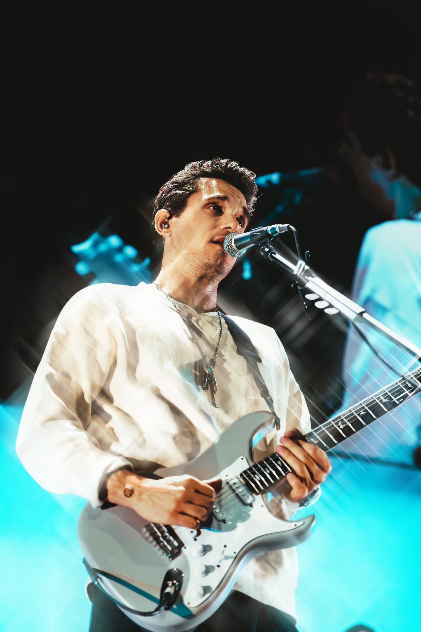 John Mayer sings into a microphone while playing a white electric guitar, backlit in pale blue.