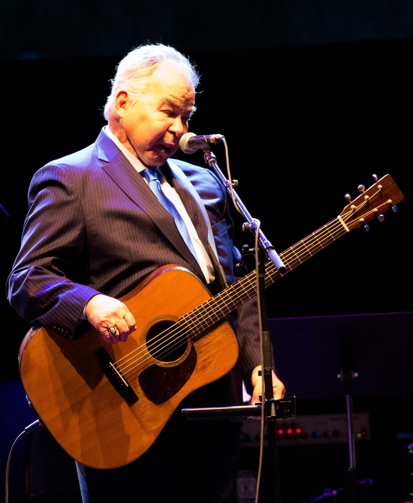 John Prine, in a pinstriped suit, sings into a microphone while playing an acoustic guitar on a dimly lit stage.