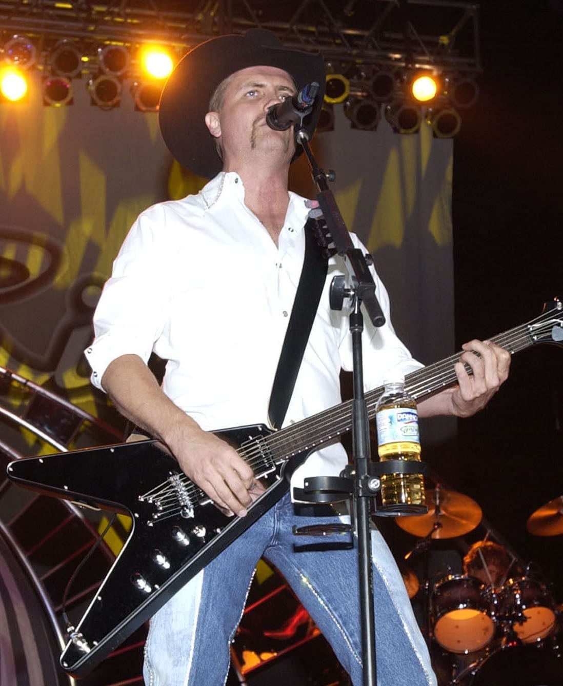 John Rich sings into a microphone on stage in a black cowboy hat and white shirt, playing a black Flying V guitar.