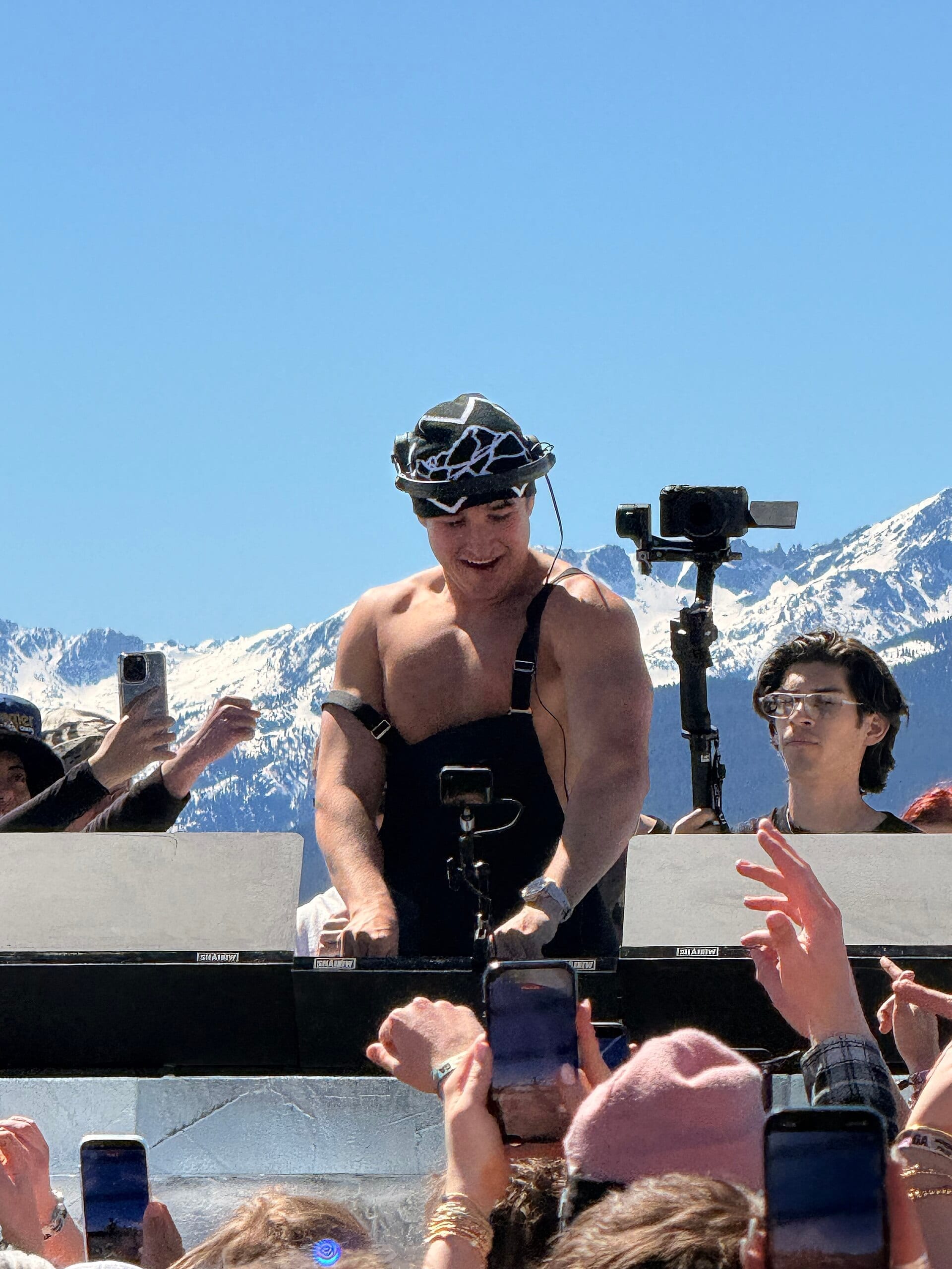 John Summit, shirtless in black overalls and a patterned hat, DJs on an outdoor stage in front of snow-capped mountains and a crowd holding up phones.