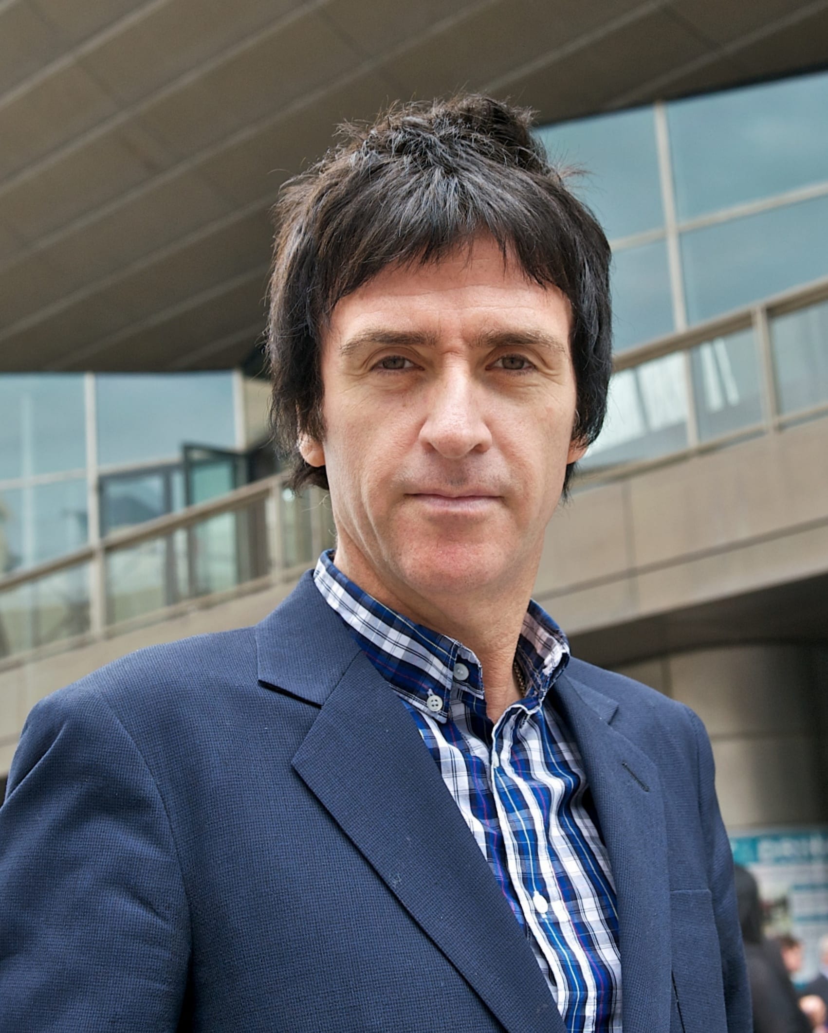 Johnny Marr, in a navy blazer and plaid shirt, looks directly at the camera outdoors.