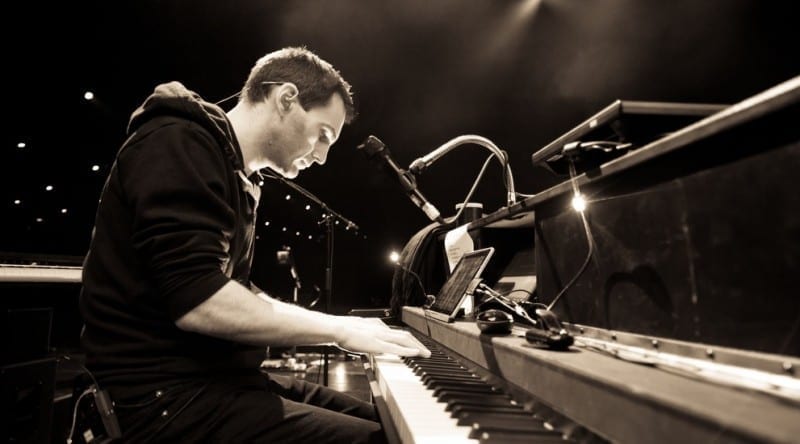 Johnny McDaid plays a grand piano on a dim stage in this sepia-toned photo, seen in profile with a microphone in front of him.