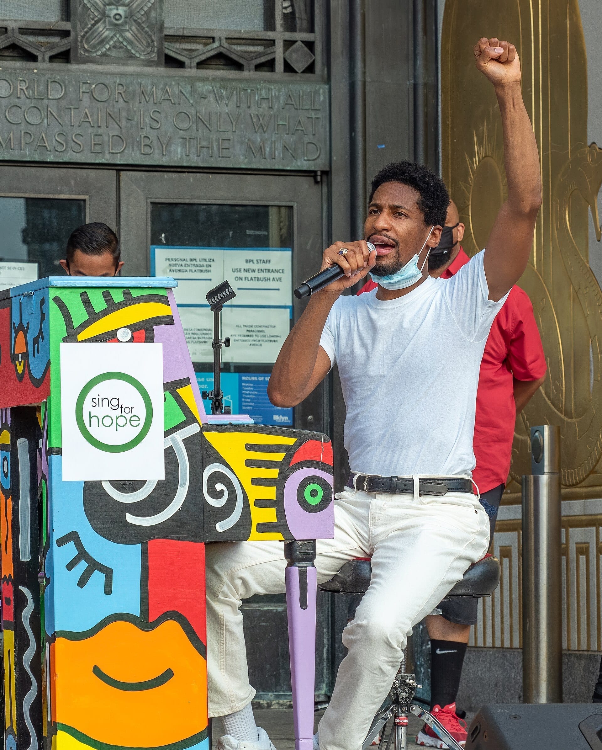 Jon Batiste sings into a microphone with his fist raised, seated at a colorfully painted piano outdoors.