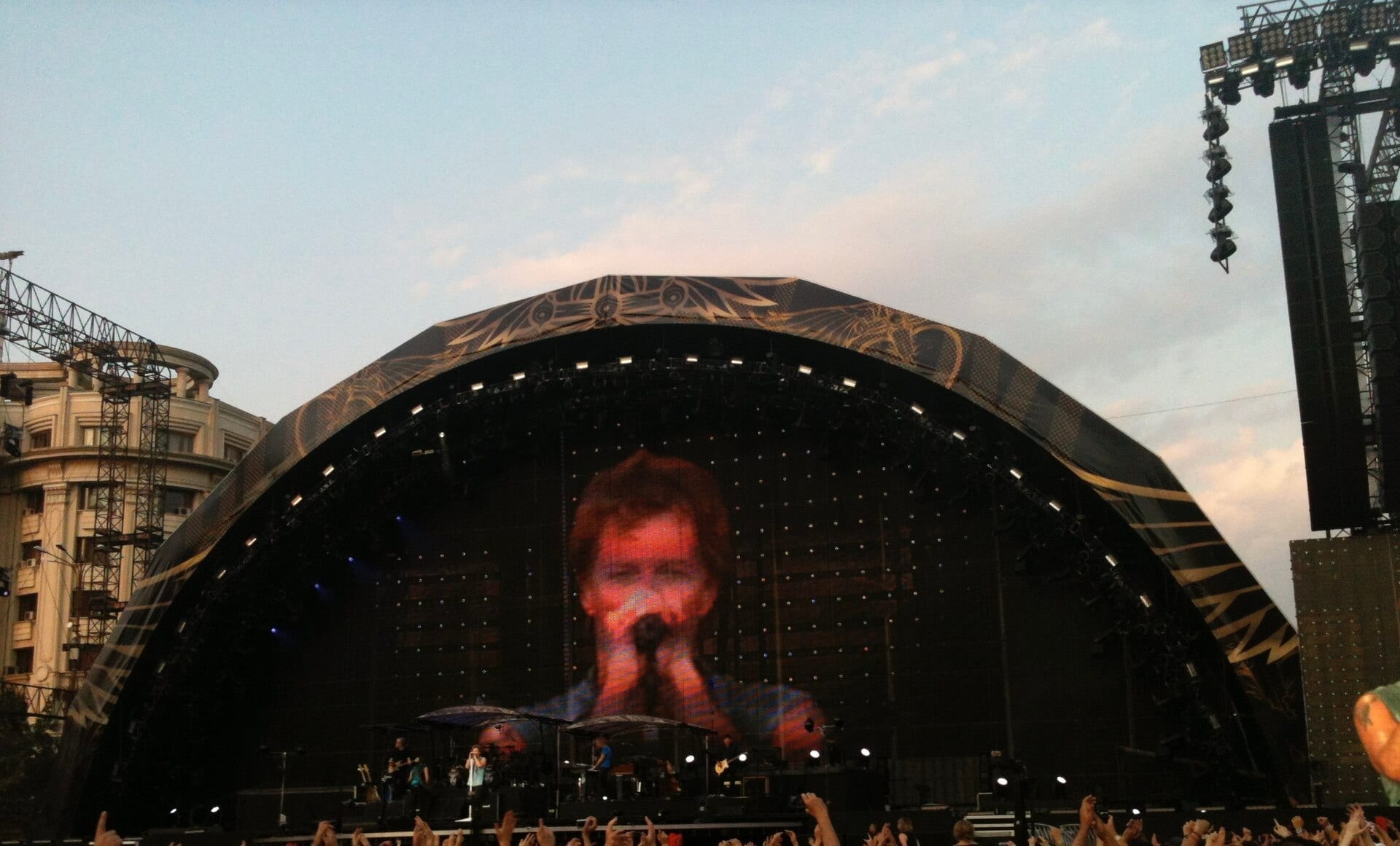 Jon Bon Jovi sings into a microphone, his face shown on a huge stage screen above the band and a cheering outdoor crowd.