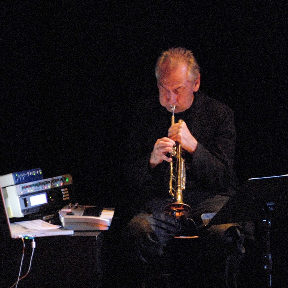 Jon Hassell, seated, plays a trumpet fitted with electronic processing gear, lit against a dark stage.
