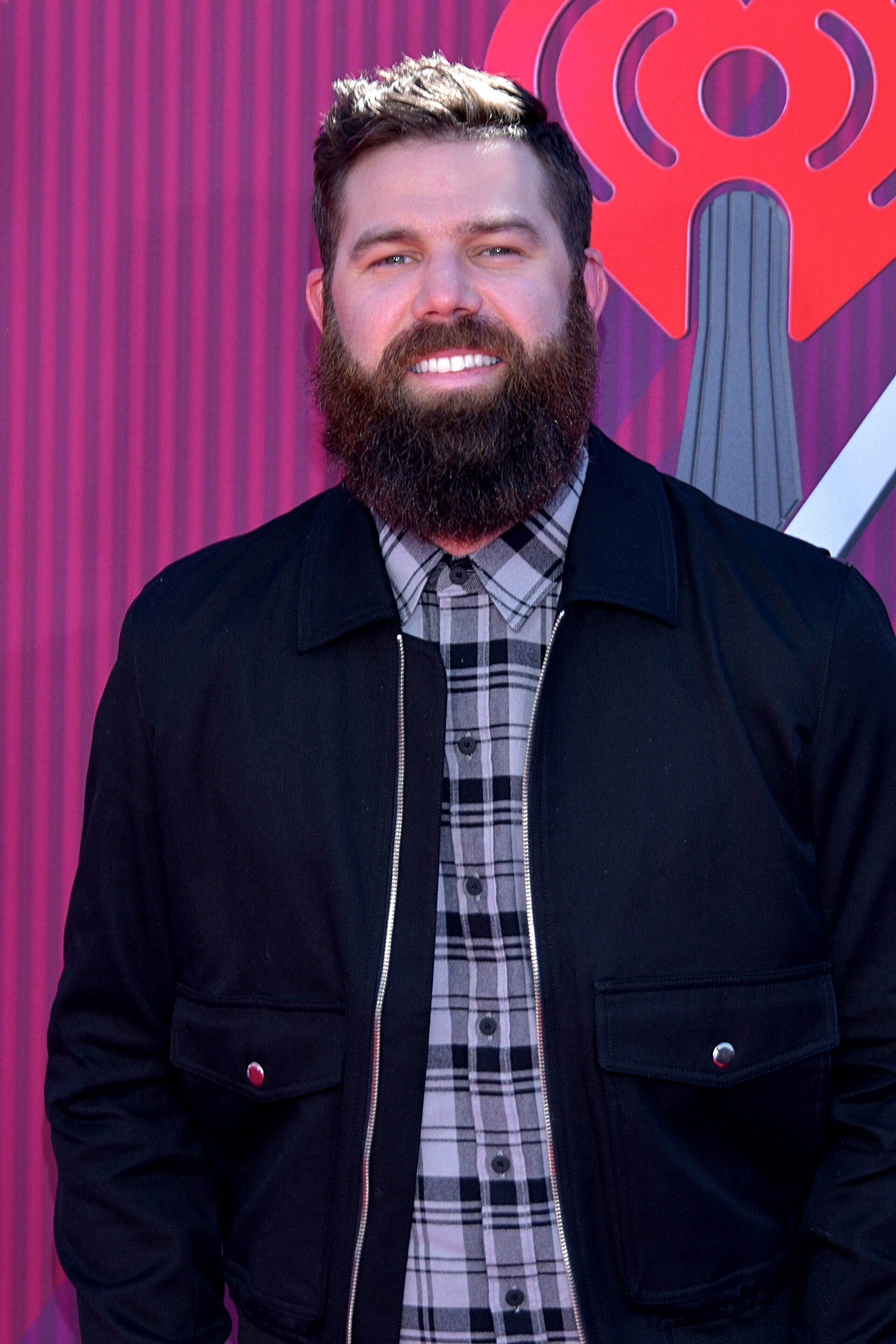 Jordan Davis, wearing a black jacket over a plaid shirt, smiles on a red carpet in front of an iHeartRadio backdrop.