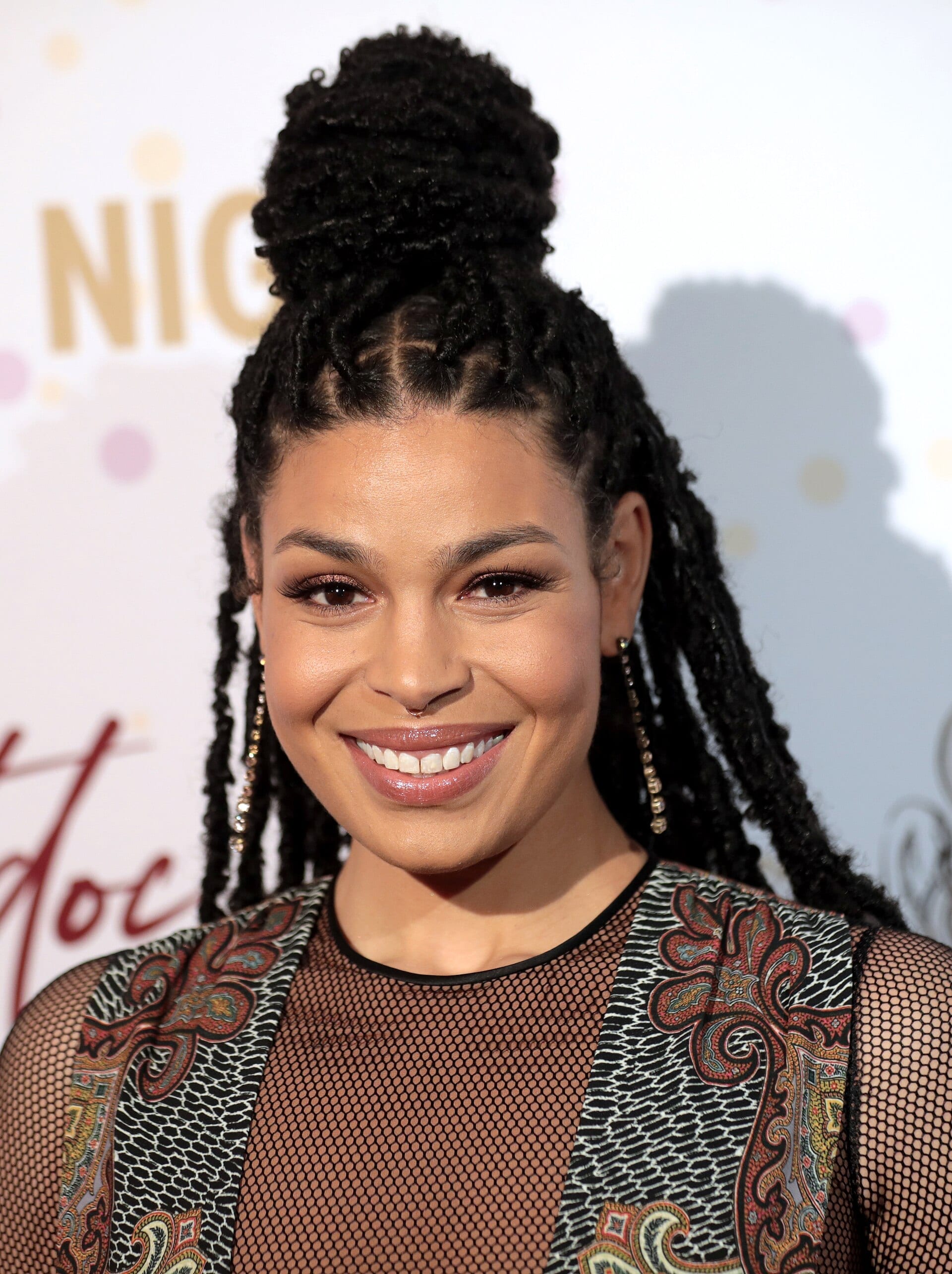 Jordin Sparks, with braided hair pulled up and long earrings, smiles at the camera in a patterned mesh top on a red carpet.